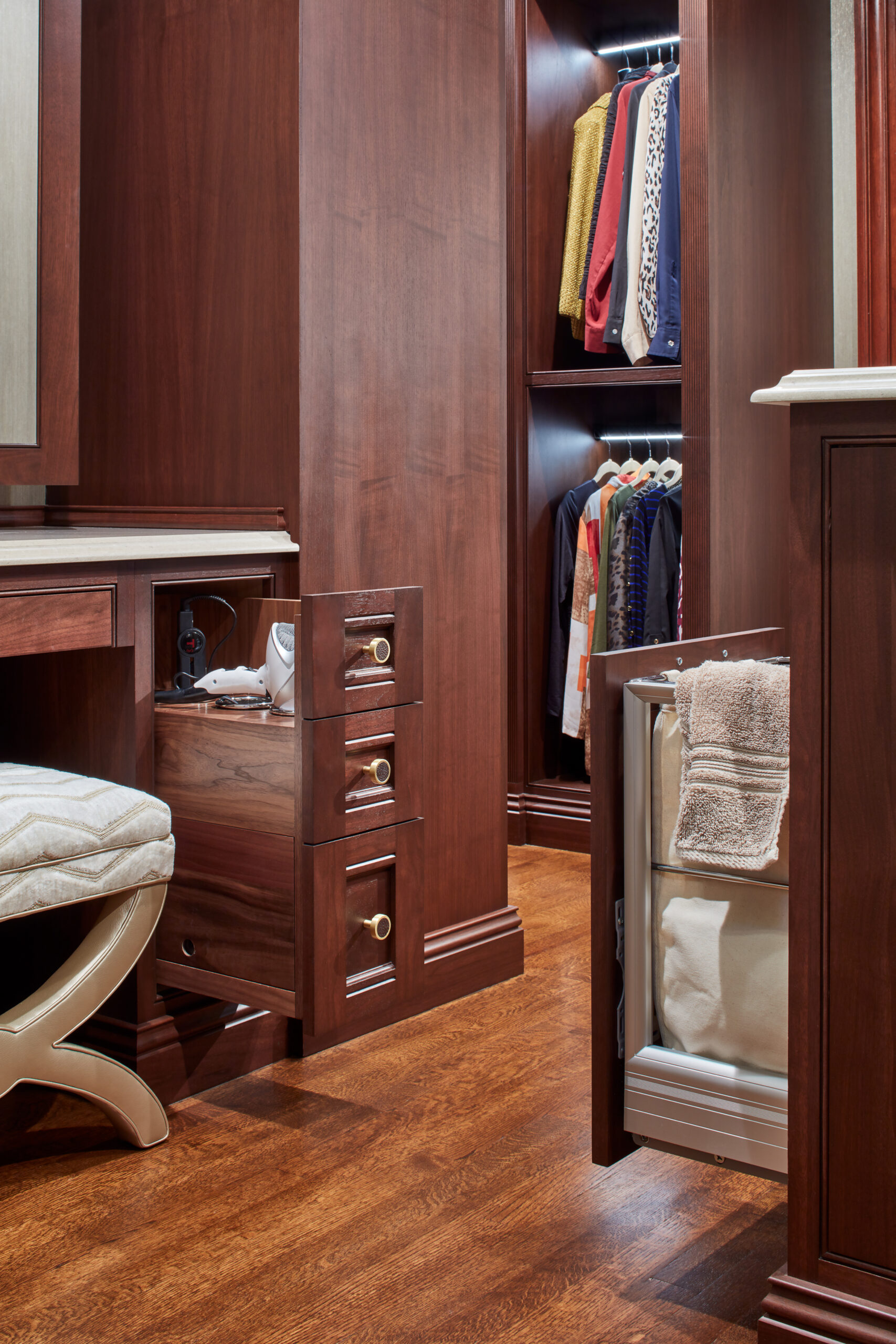 A walk-in closet with wooden cabinetry, an open drawer, an open built-in laundry hamper, hanging clothes, and a cushioned vanity stool on a wood floor exemplifies luxury interior design by an expert Interior Designer Massachusetts.