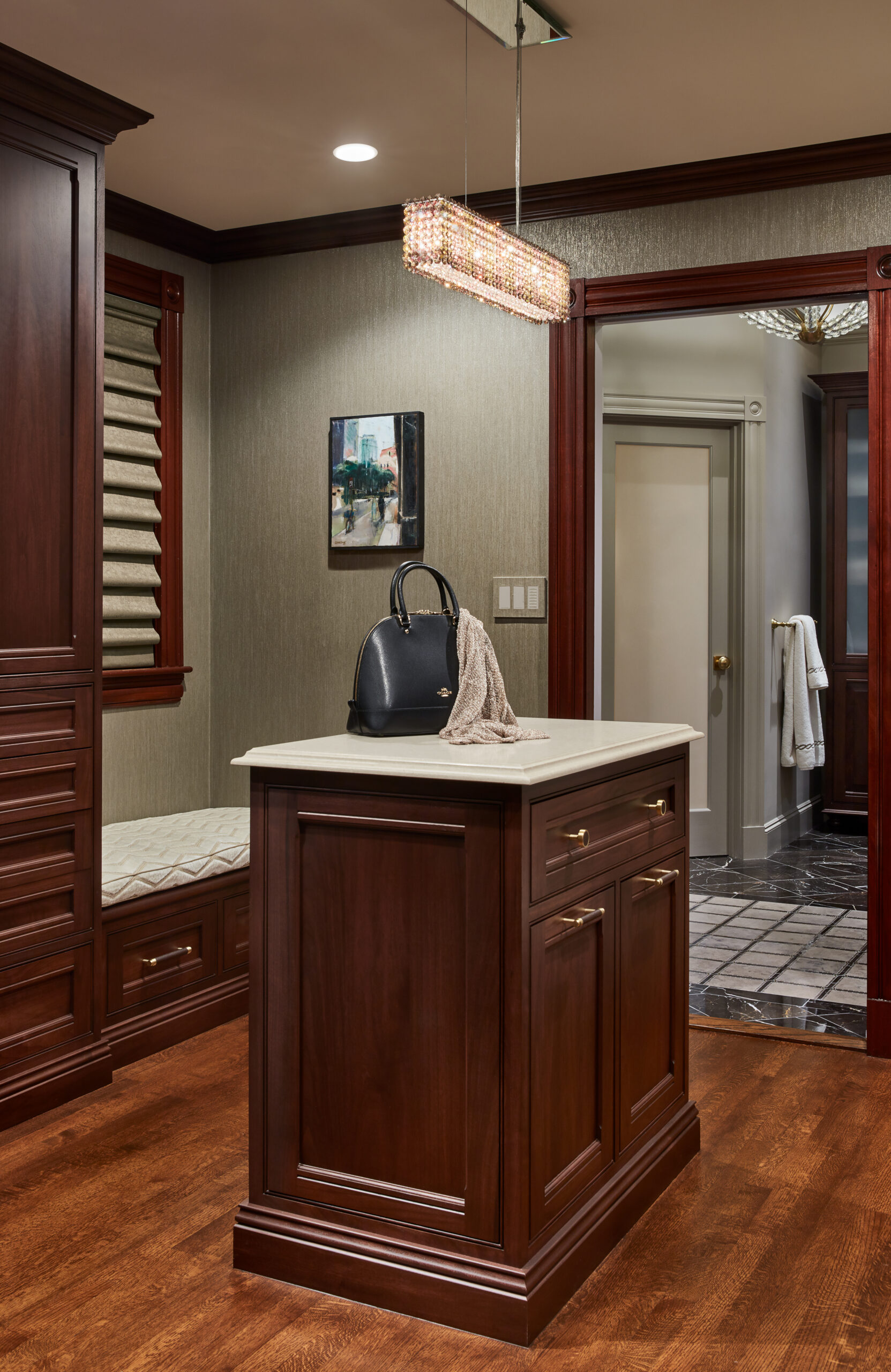 A small room with dark wood cabinetry, a central island, a black handbag and scarf, bench seating, framed art, and a doorway leading to a tiled bathroom—crafted with the elegance typical of luxury interior design.