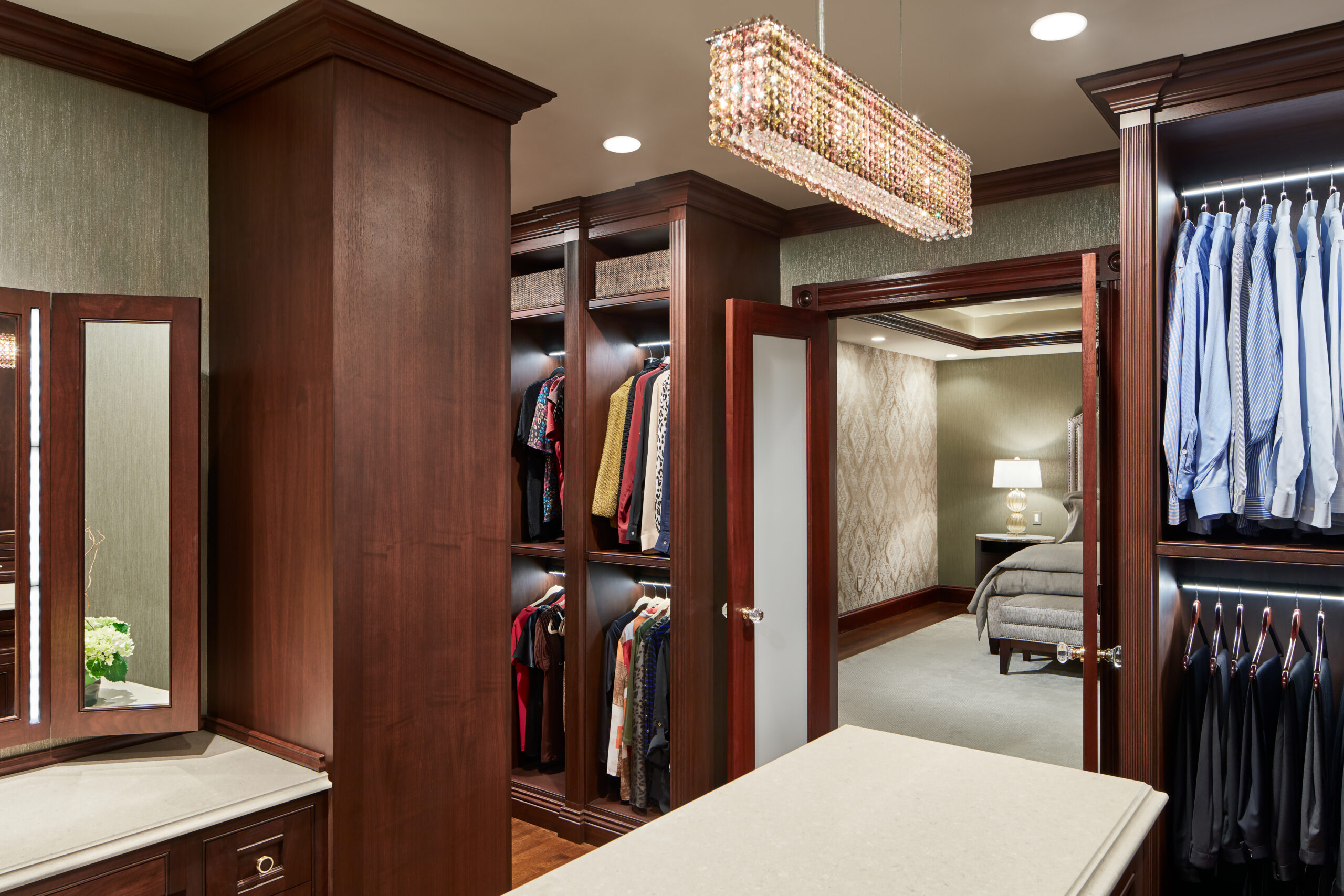 A walk-in closet with wooden shelves and hanging clothes, a vanity with a mirror, and a view into a bedroom with a bed and nightstand—reflecting the elegance of Luxury Interior Design.