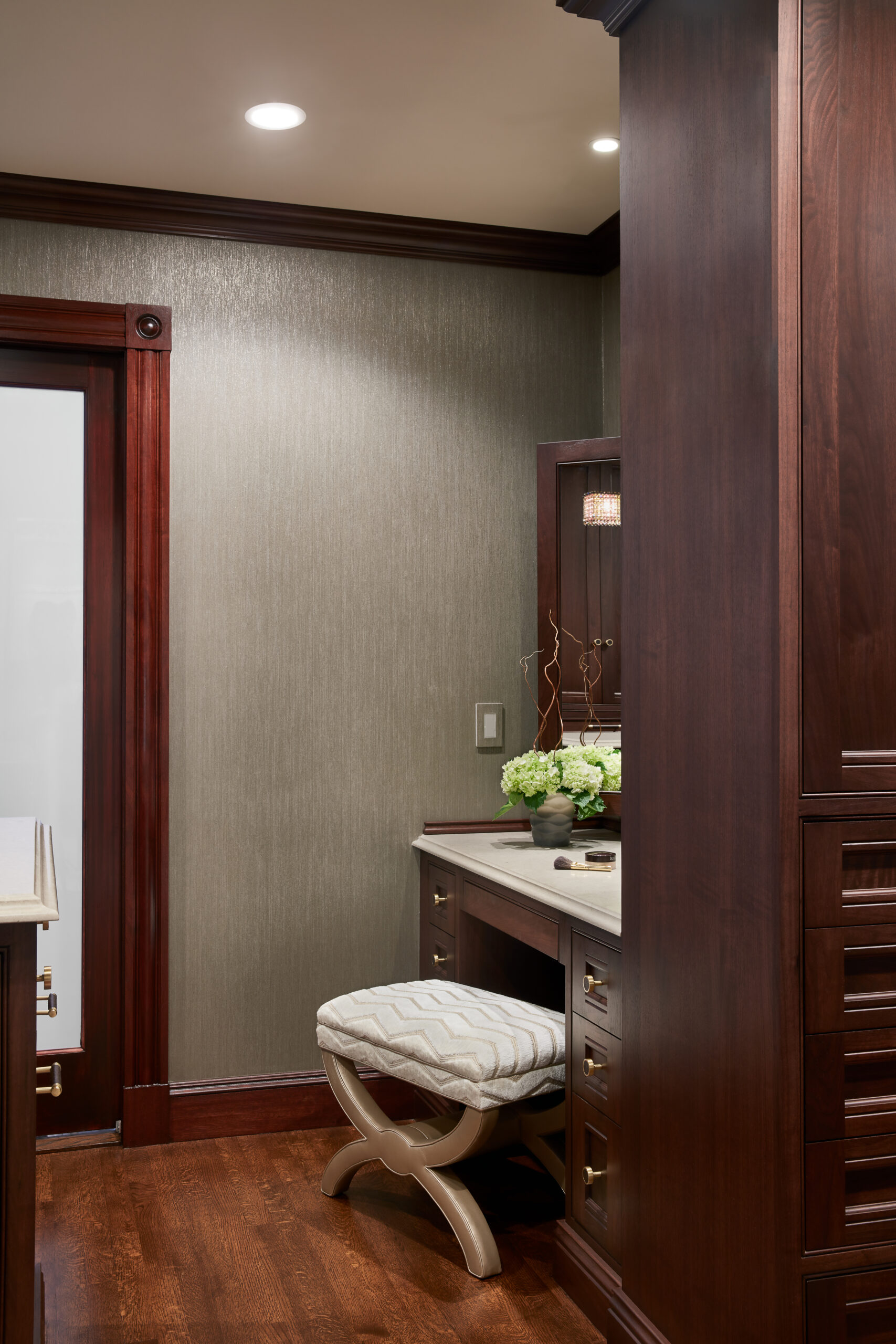 A small vanity area with luxury interior design, featuring a cushioned stool, dark wood cabinetry, a countertop, a vase of flowers, and a textured gray wall.