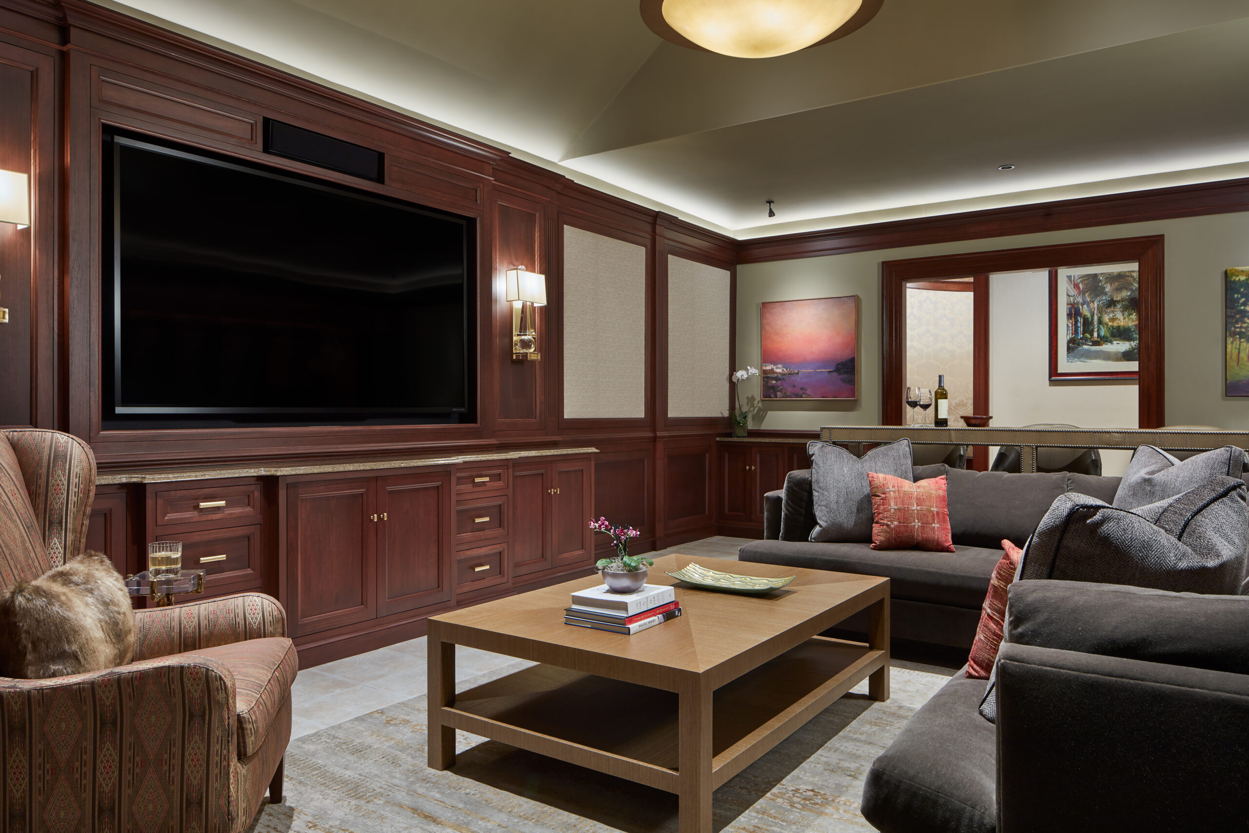 A living room with dark wood paneling, a large wall-mounted TV, gray sofa, armchair, wooden coffee table, and artwork on the walls showcases Luxury Interior Design perfect for a sophisticated Massachusetts home.