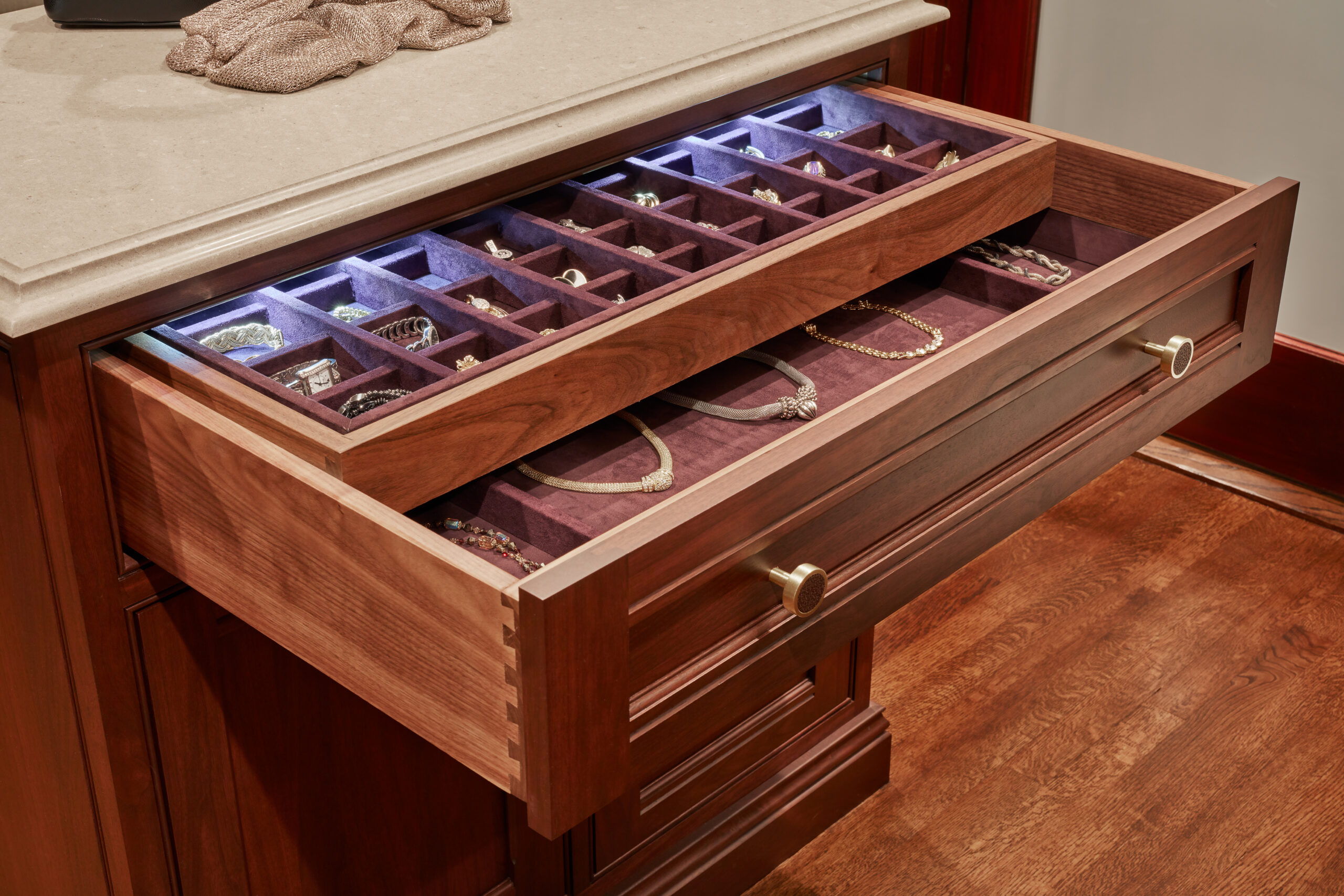 A wooden drawer with purple velvet-lined compartments displays organized jewelry—rings, bracelets, and necklaces—beneath a countertop, embodying the elegance of luxury interior design.