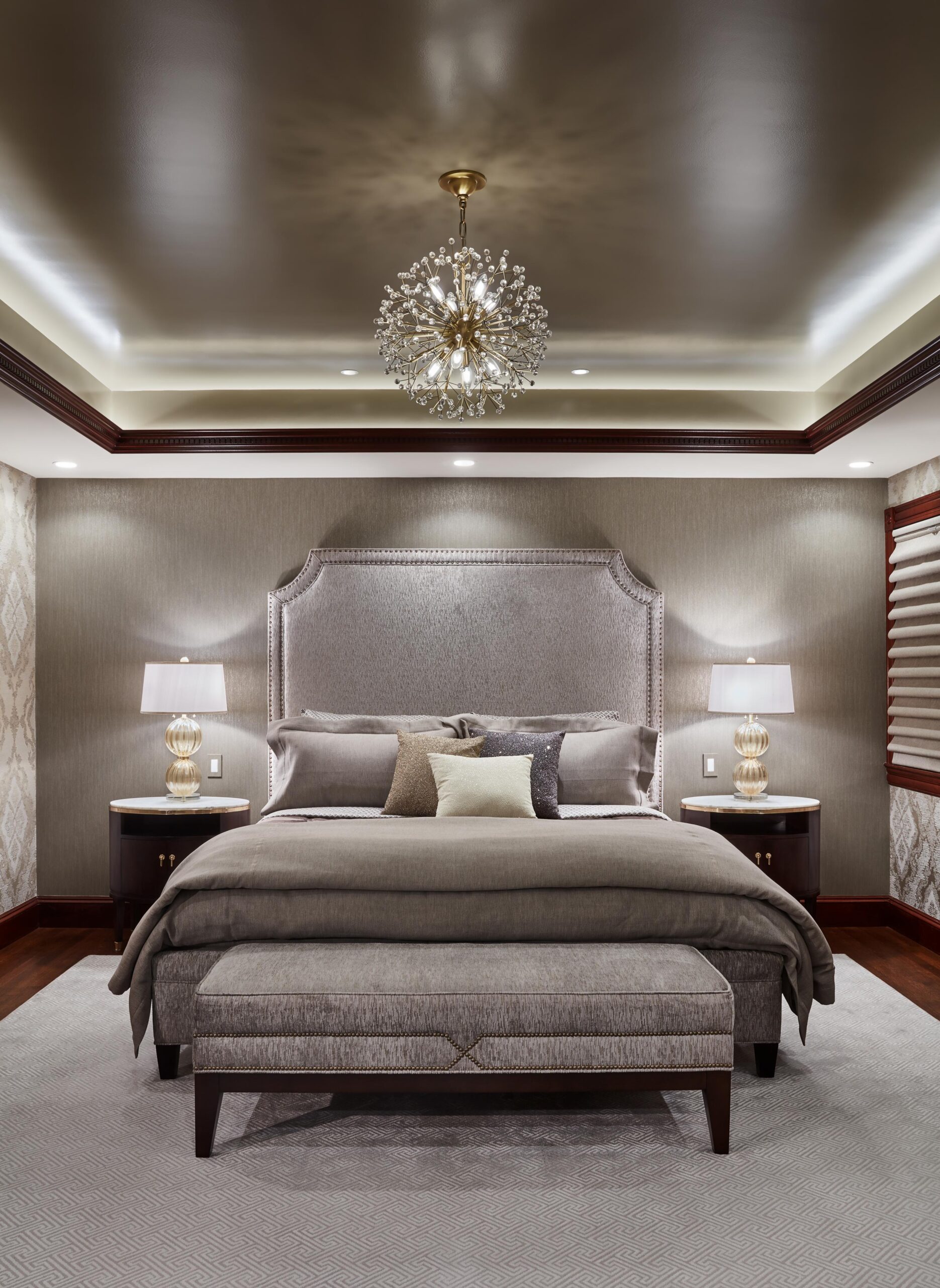 Elegant bedroom living space featuring a large upholstered bed, two bedside tables with lamps, a bench at the foot of the bed, and a modern chandelier centered on the ceiling.