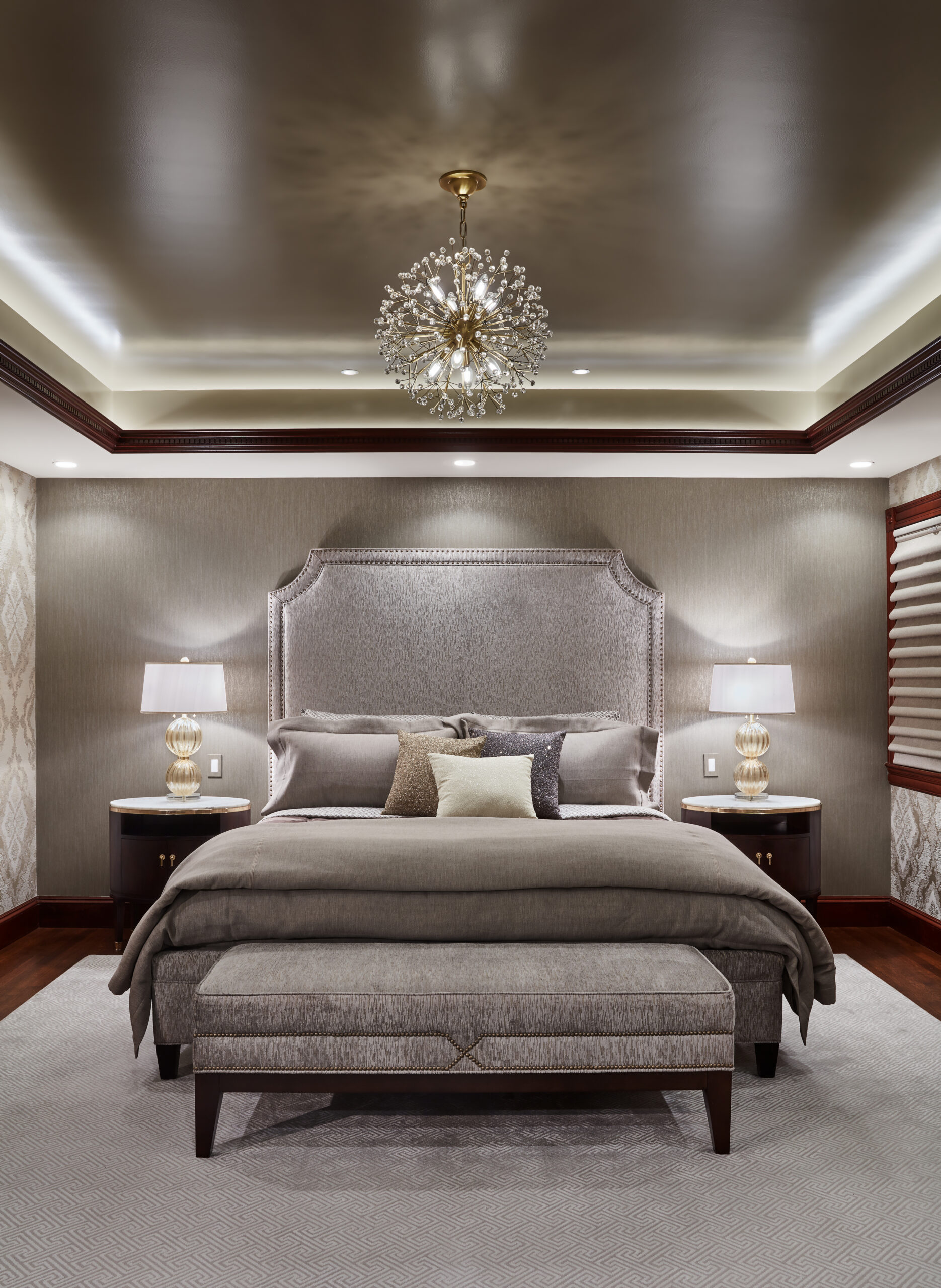 Elegant bedroom with a large upholstered bed, bench at the foot, two nightstands with lamps, neutral decor, and a chandelier on a tray ceiling—showcasing classic Luxury Interior Design by an expert interior designer in Massachusetts.
