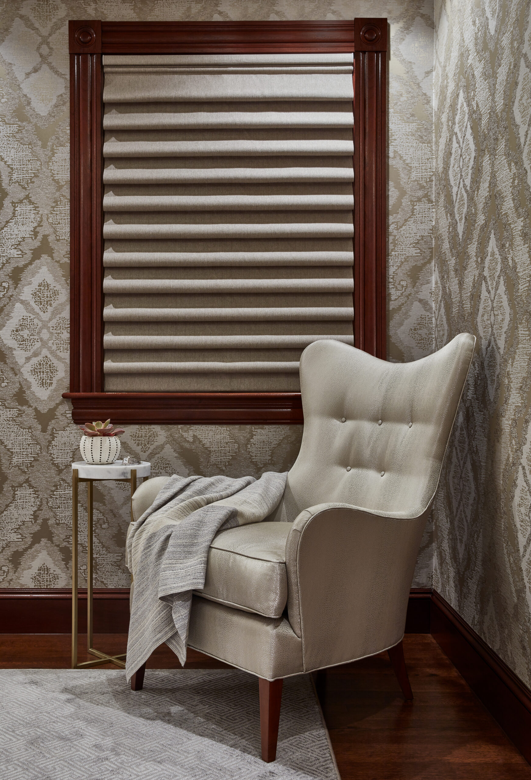 Beige armchair with a blanket draped over the arm sits next to a small table with a decorative item, beneath a window with a closed fabric shade in a wallpapered room—perfect for those seeking luxury interior design inspiration.