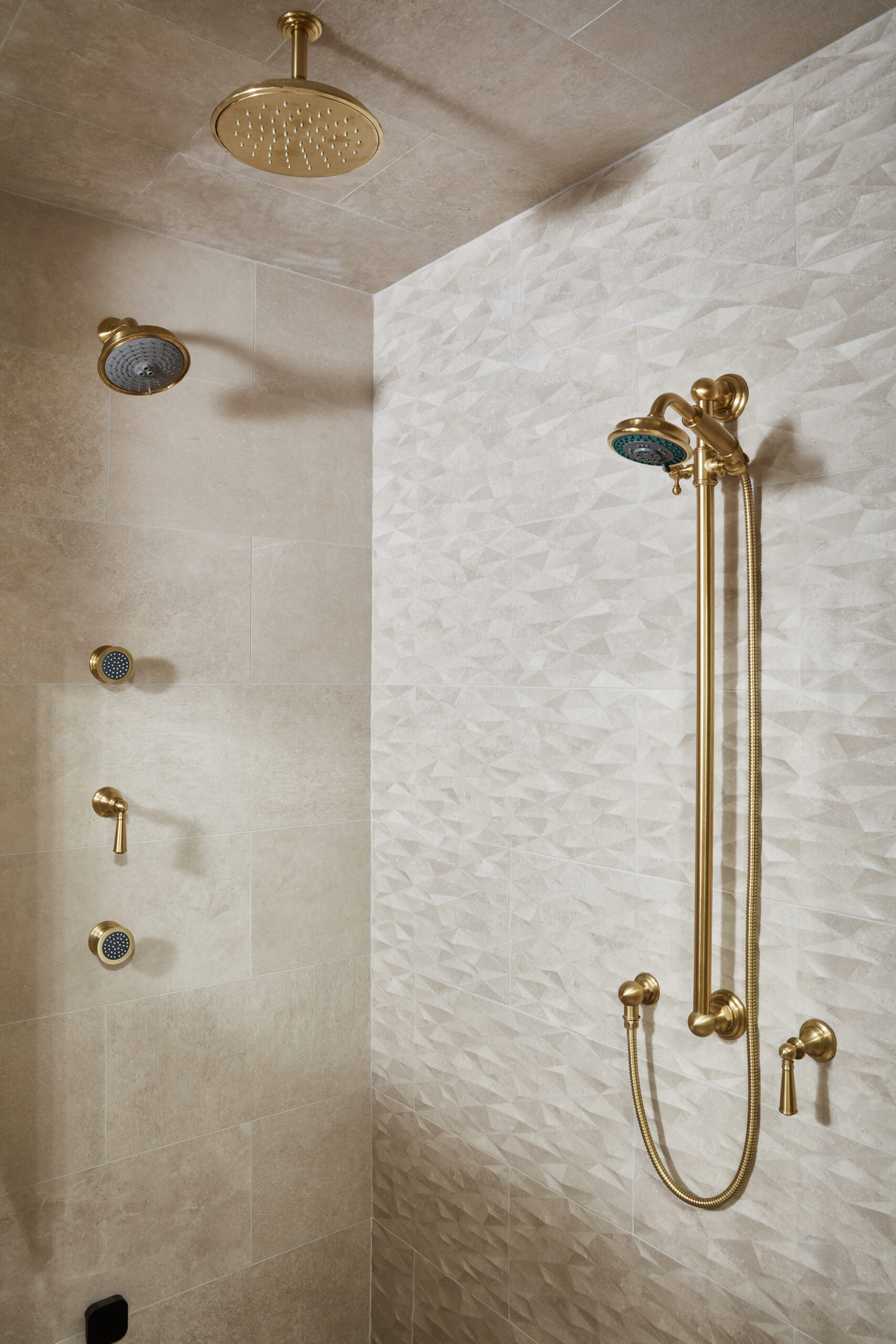 A modern shower with gold fixtures, including a rainfall showerhead, wall-mounted hand shower, and textured beige tile walls—perfect for those seeking luxury interior design by an expert interior designer in Massachusetts.