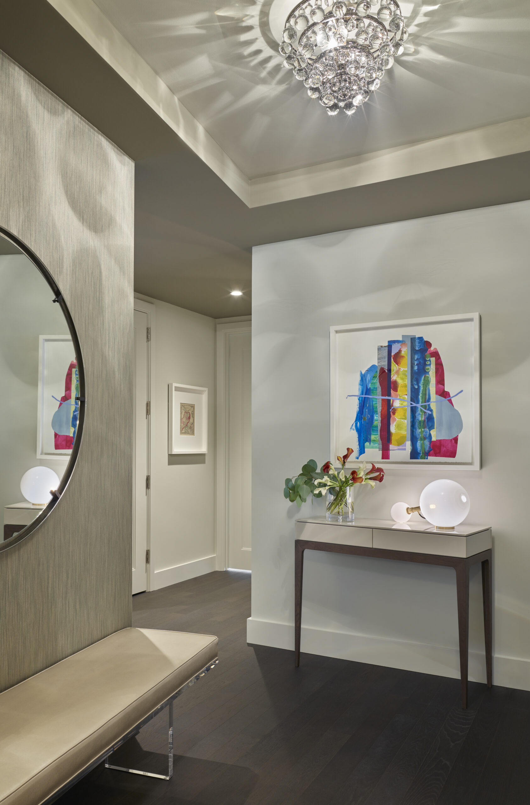 Modern entryway with dark wood floors, a bench, round mirror, console table with decor items, abstract artwork on the wall, and a crystal chandelier—perfectly capturing Luxury Interior Design by an expert Interior Designer Massachusetts.