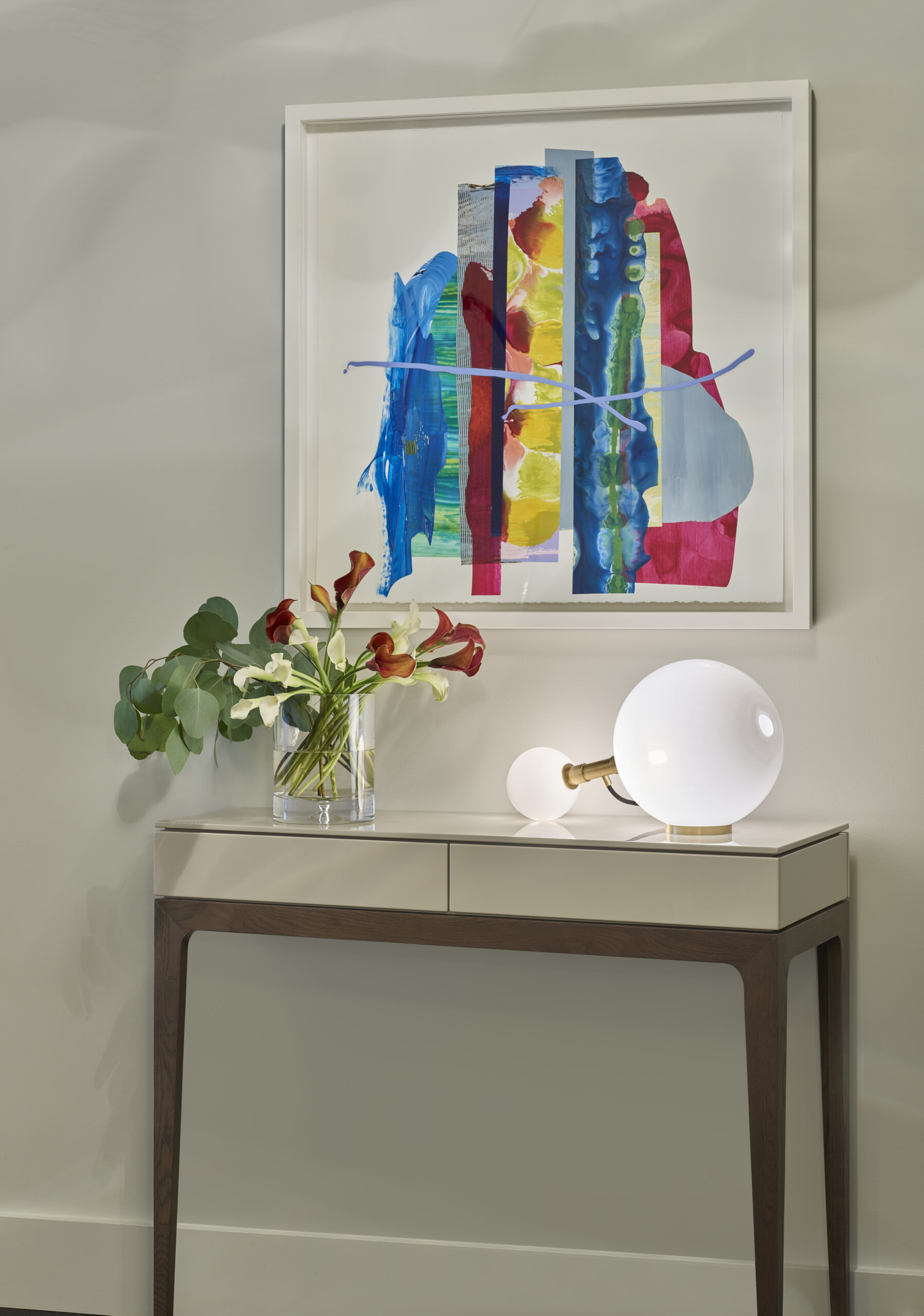 A modern console table with a vase of flowers and a spherical lamp, beneath a colorful abstract painting on a light-colored wall, showcases the refined touch of luxury interior design by an expert interior designer in Massachusetts.
