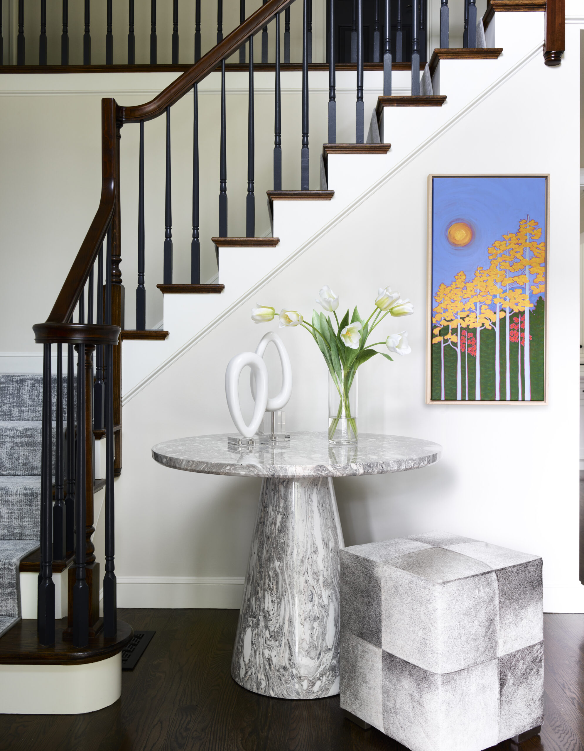 A marble table with a glass vase of flowers and abstract sculpture sits below a staircase; a colorful painting and cube-shaped stool nearby add to the Luxury Interior Design crafted by an expert Interior Designer Massachusetts.