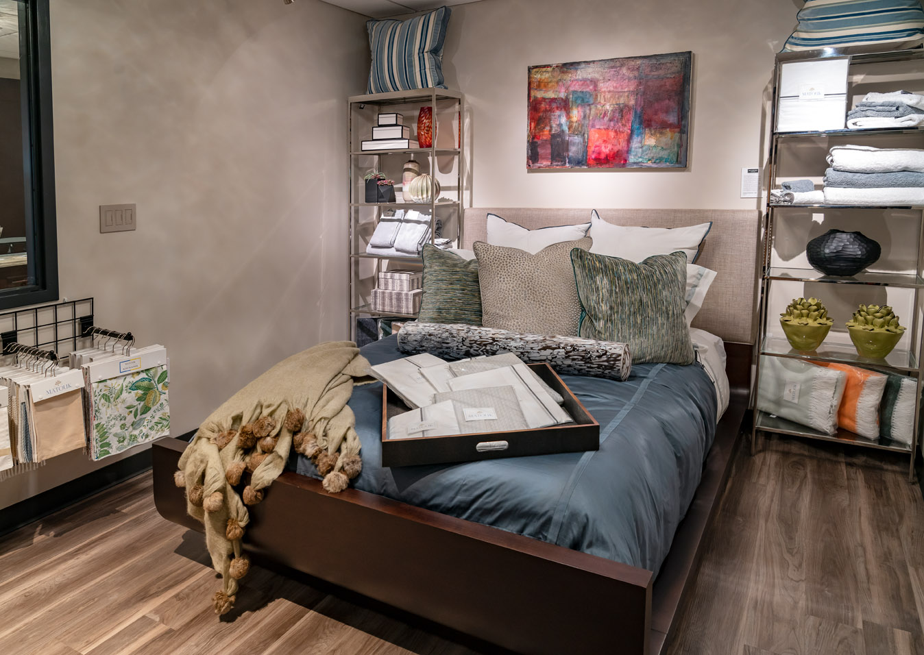 A neatly made bed with assorted pillows and a throw blanket anchors this modern bedroom, surrounded by shelves with towels, linens, and decor—perfect inspiration from the Experience Studio.
