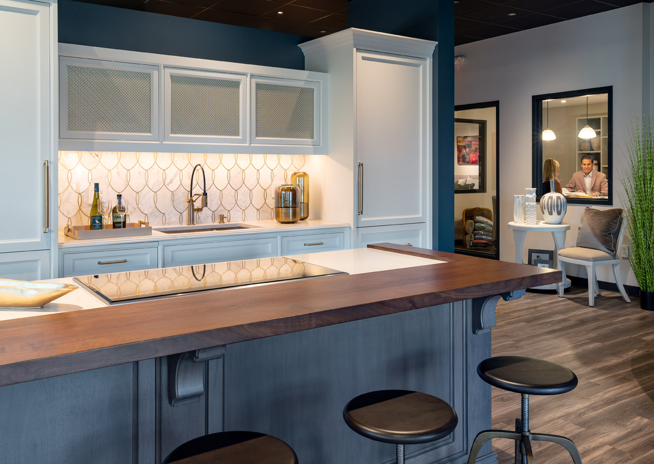 Modern kitchen with white cabinets, a wooden island countertop, bar stools, and geometric tile backsplash; two people converse in a separate glass-walled Experience Studio in the background.