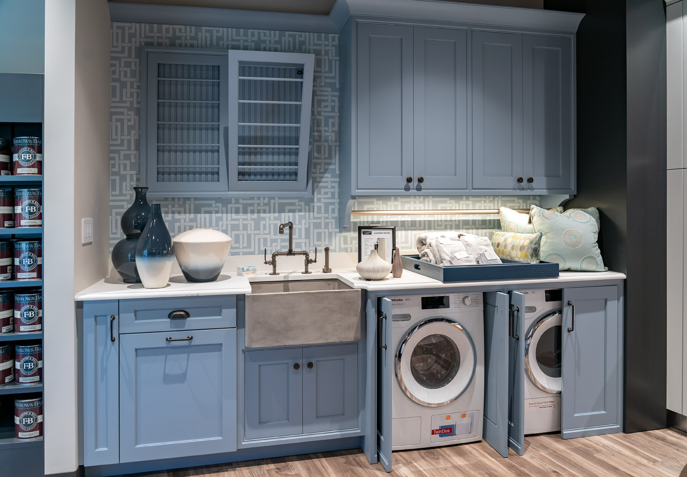 Modern laundry room with blue cabinets, farmhouse sink, washer and dryer, countertop decor, and patterned backsplash. Cushions and a tray are placed on the counter above the appliances for a cozy Experience Studio touch.