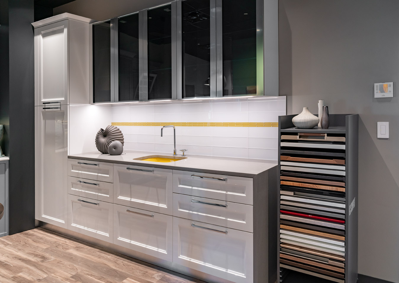 Modern kitchen with glossy white cabinets, glass-front uppers, a yellow-accented backsplash, and a display rack of cabinet door samples on the right—perfect for exploring options in this Experience Studio.