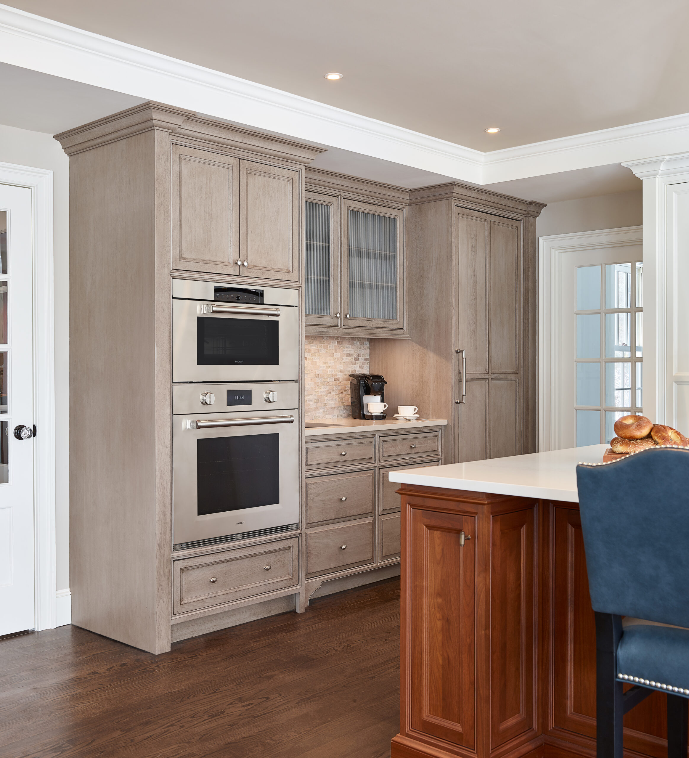 Modern kitchen with light wood cabinets, built-in double ovens, glass-fronted uppers, tile backsplash, and a white island with dark wood base and blue chair—an elegant example of luxury interior design by an Interior Designer Massachusetts.