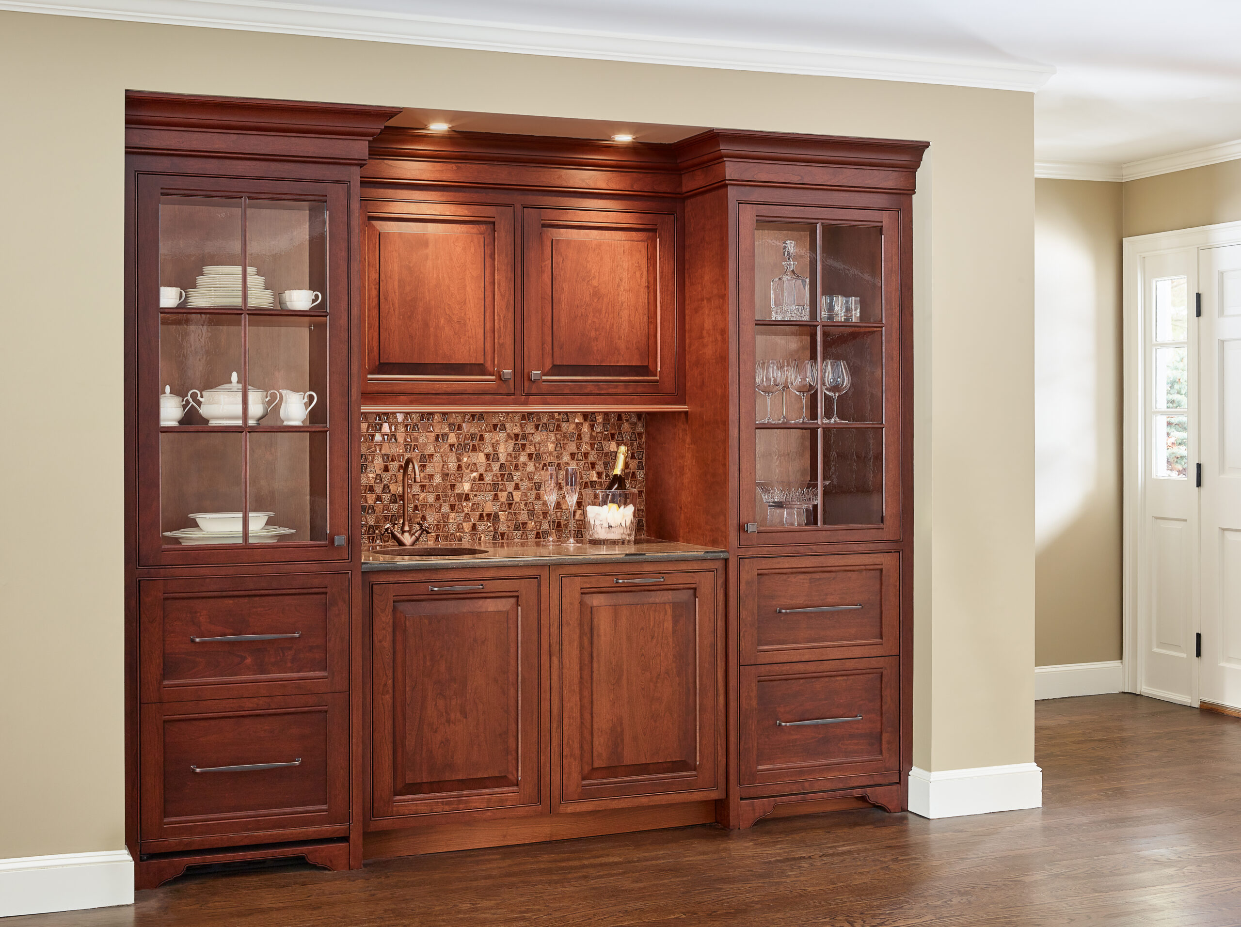 A built-in wooden cabinet with glass doors displays dishes and glassware, featuring a mosaic tile backsplash and under-cabinet lighting. Designed by a top Interior Designer Massachusetts, it brings luxury interior design to your home.