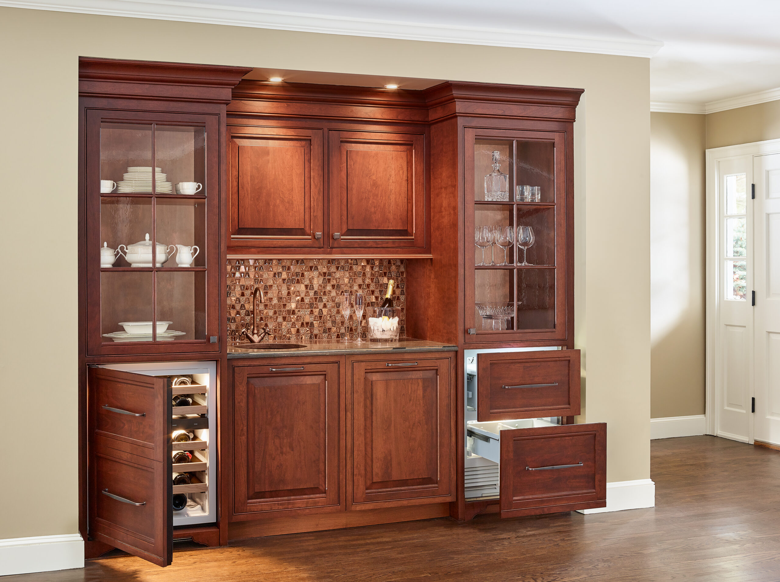 A wooden kitchen cabinet with glass doors displays dishes and glasses; lower drawers reveal wine storage—an elegant touch that reflects luxury interior design curated by an expert interior designer in Massachusetts.