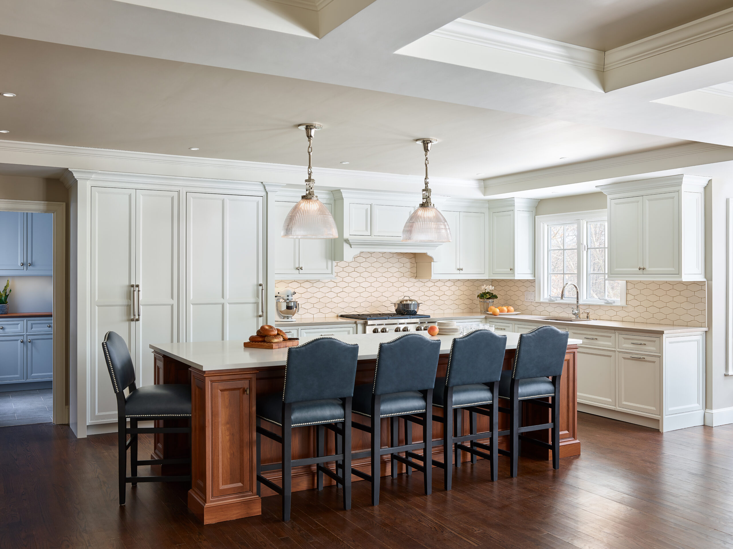 Spacious kitchen with white cabinets, a large island with dark chairs, pendant lights, and wood flooring. Designed by an Interior Designer Massachusetts, natural light enters through a window above the sink for a touch of luxury interior design.