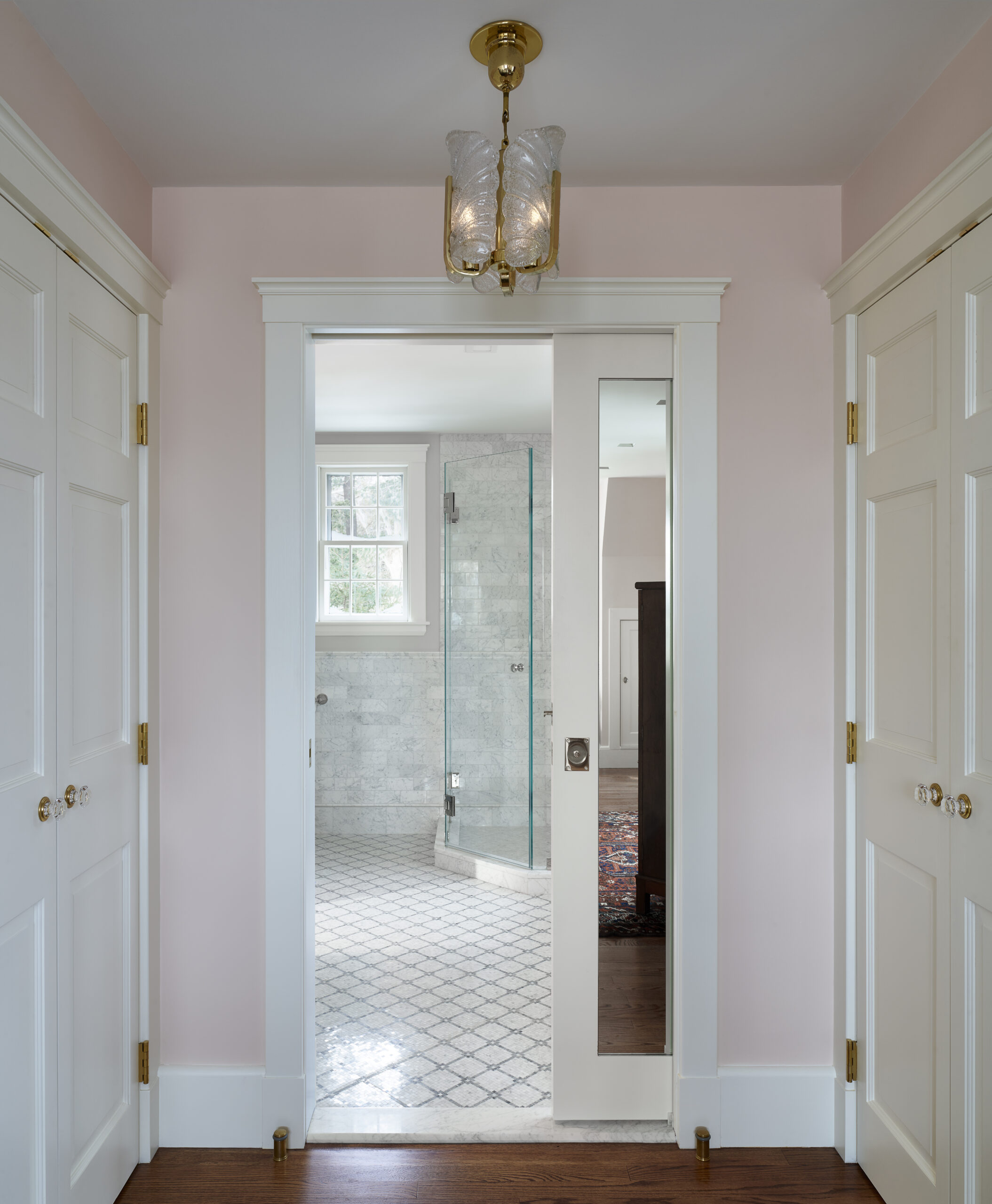 Hallway with white double doors leads to a bathroom designed by an Interior Designer Massachusetts, featuring a glass shower, patterned tile floor, a window, and a gold light fixture—a perfect touch of luxury interior design.