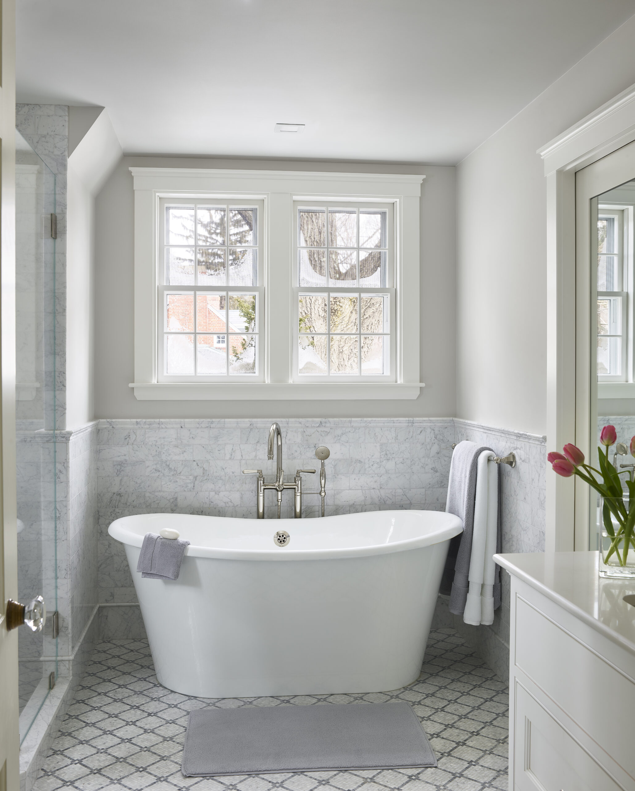 Bright bathroom with a freestanding bathtub, gray mat, towel, marble walls, two windows, and a vase of pink tulips on a white countertop—perfect inspiration for luxury interior design by an Interior Designer Massachusetts.