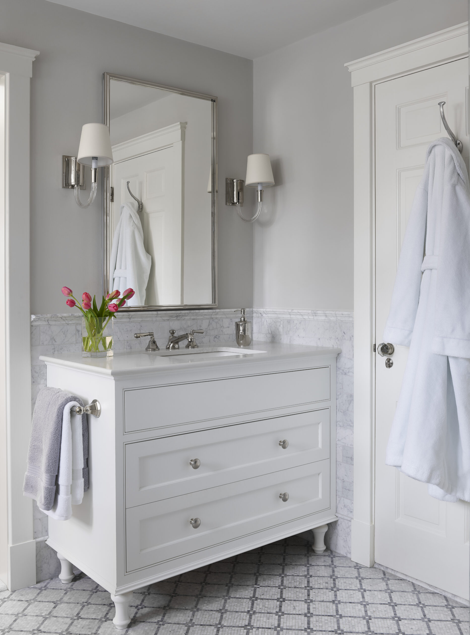 A white bathroom vanity with a marble countertop, silver fixtures, a mirror, wall sconces, a vase of red tulips, and a robe hanging on the door creates a look worthy of Luxury Interior Design by an Interior Designer in Massachusetts.