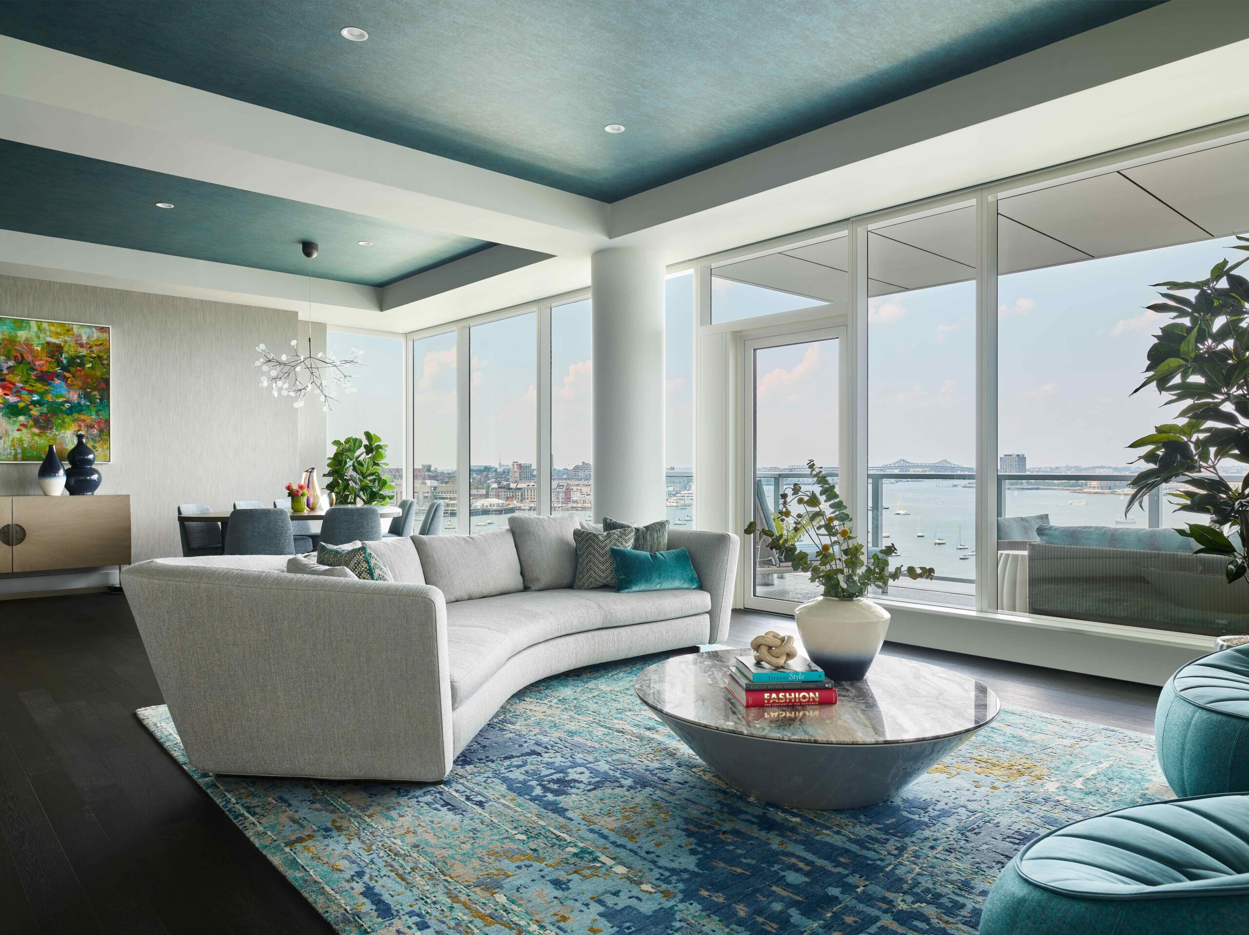 Modern living room with large windows, gray curved sofa, round marble coffee table, blue patterned rug, plants, and cityscape view outside—perfectly embodying luxury interior design.