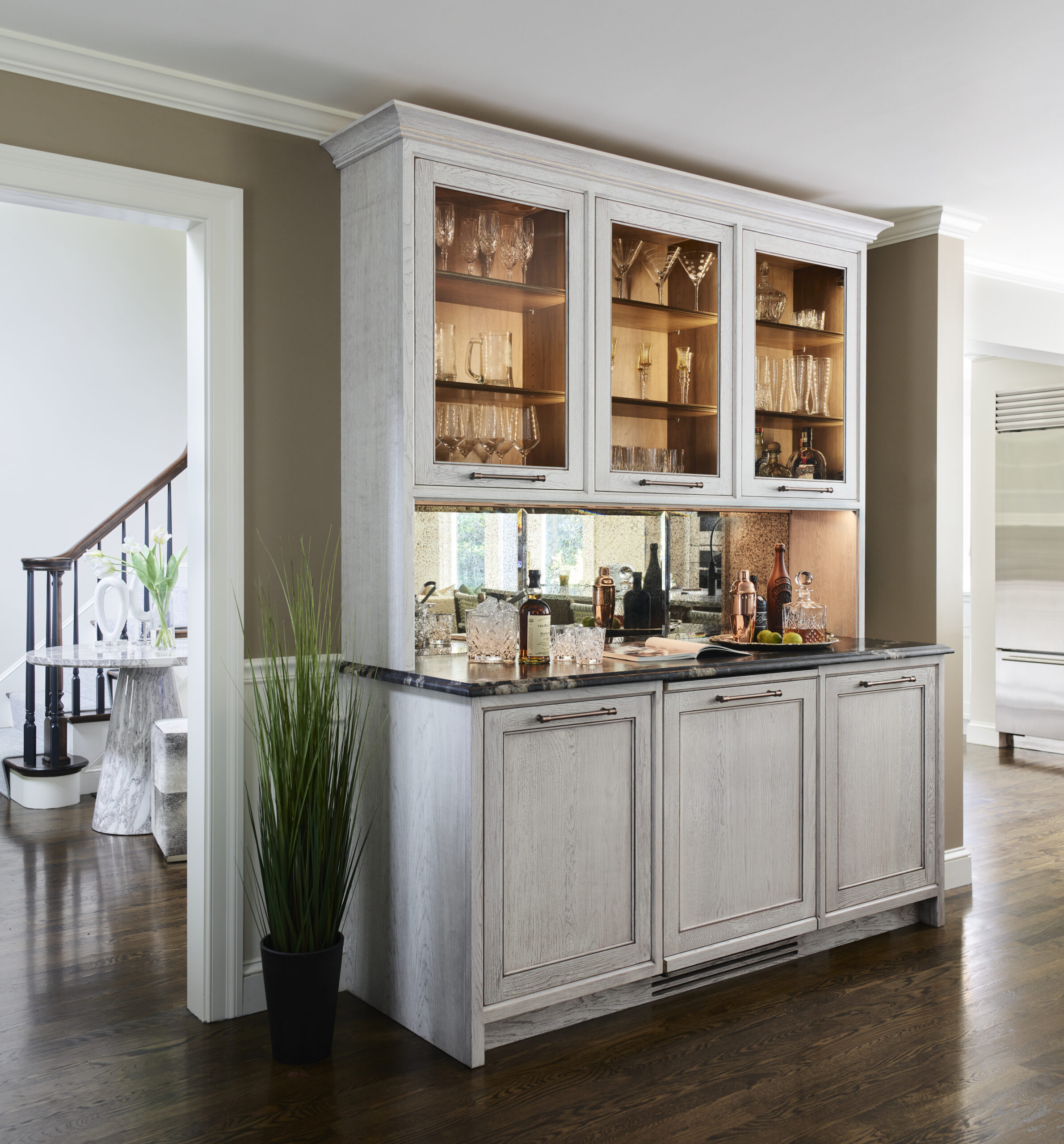 A built-in home bar with glass-front upper cabinets displays glassware, while a countertop holds bottles and accessories—reflecting the touch of a skilled Interior Designer Massachusetts. A tall plant adds greenery to this luxury interior design.