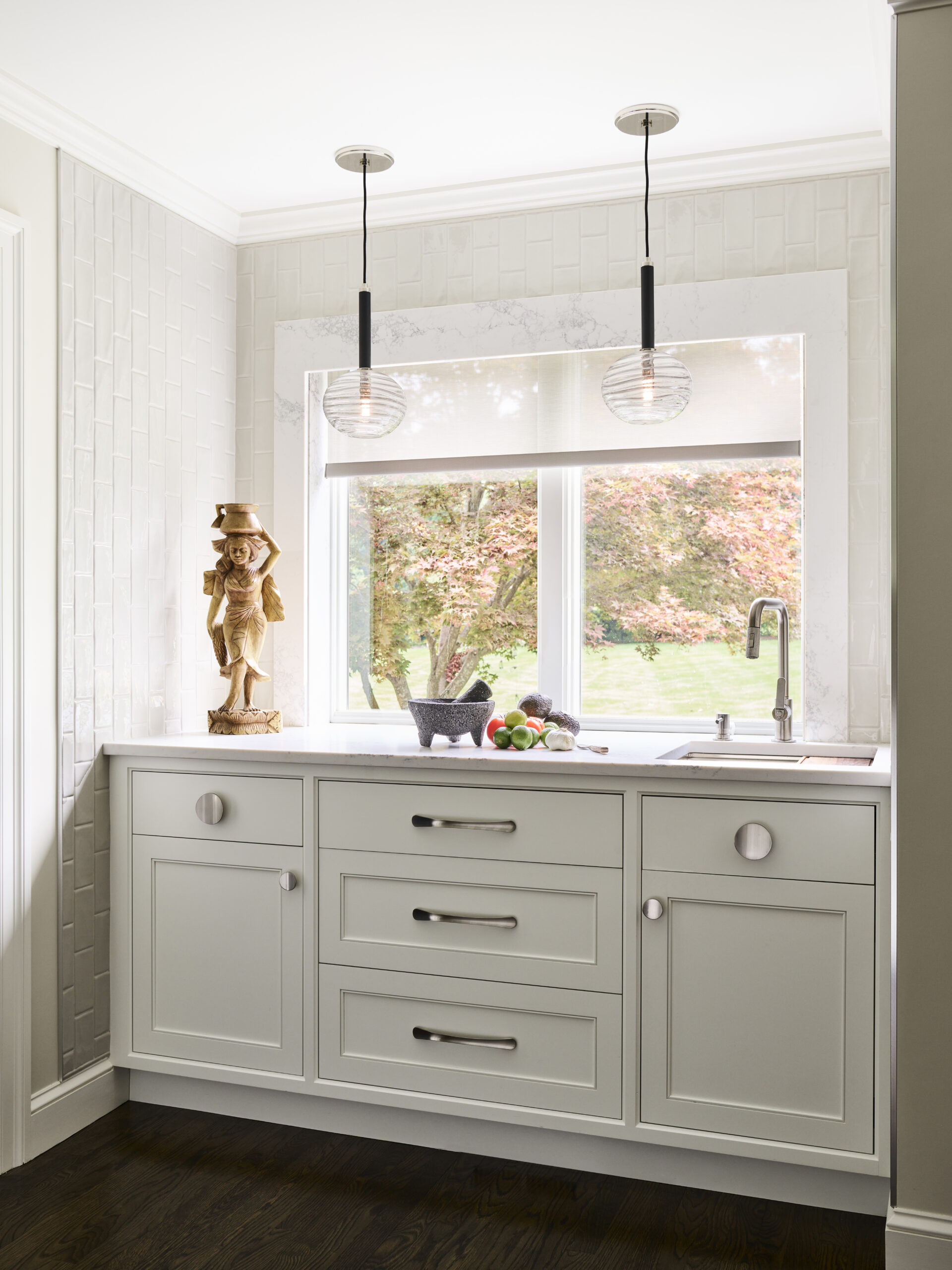 A bright kitchen nook with a white sink, gray cabinets, hanging pendant lights, a wood statuette, and a window overlooking a leafy yard—showcasing luxury interior design by an expert Interior Designer in Massachusetts.