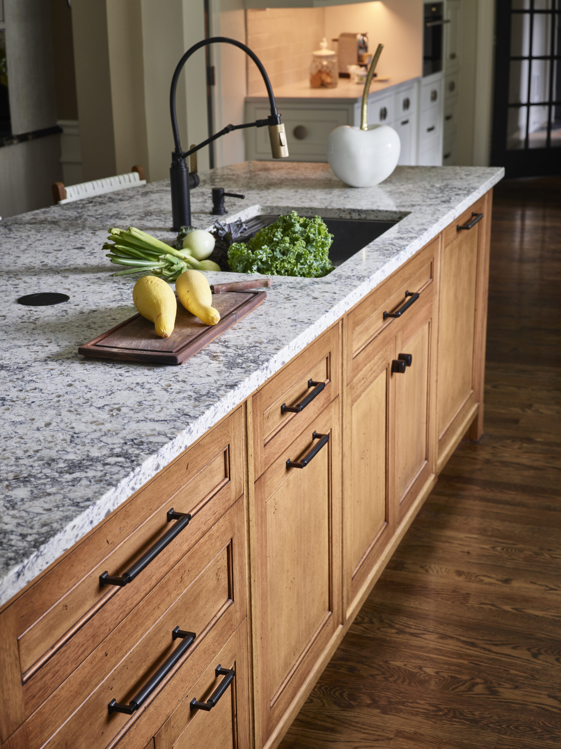 A kitchen island with wooden cabinets, a granite countertop, and a black sink showcases fresh vegetables beside a cutting board—a perfect example of Luxury Interior Design by an expert Interior Designer in Massachusetts.