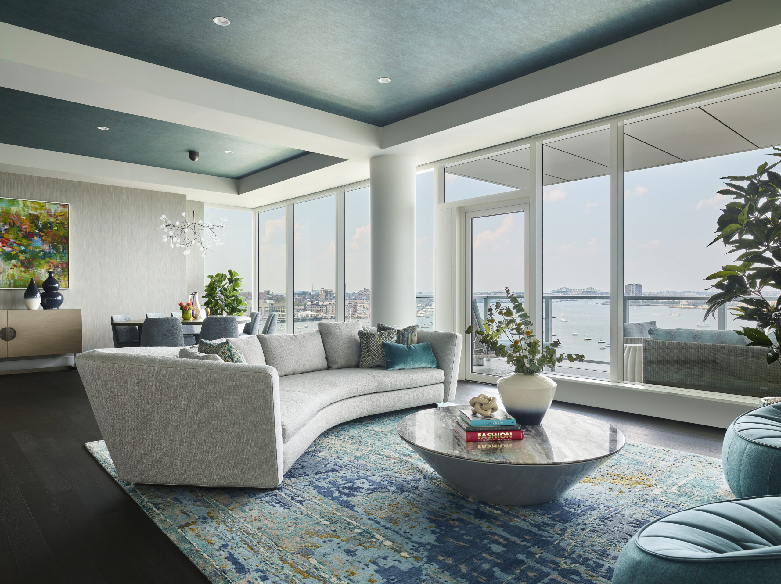 Modern living room with curved gray sofa, marble coffee table, large windows overlooking water, blue patterned rug, and contemporary décor showcasing luxury interior design.