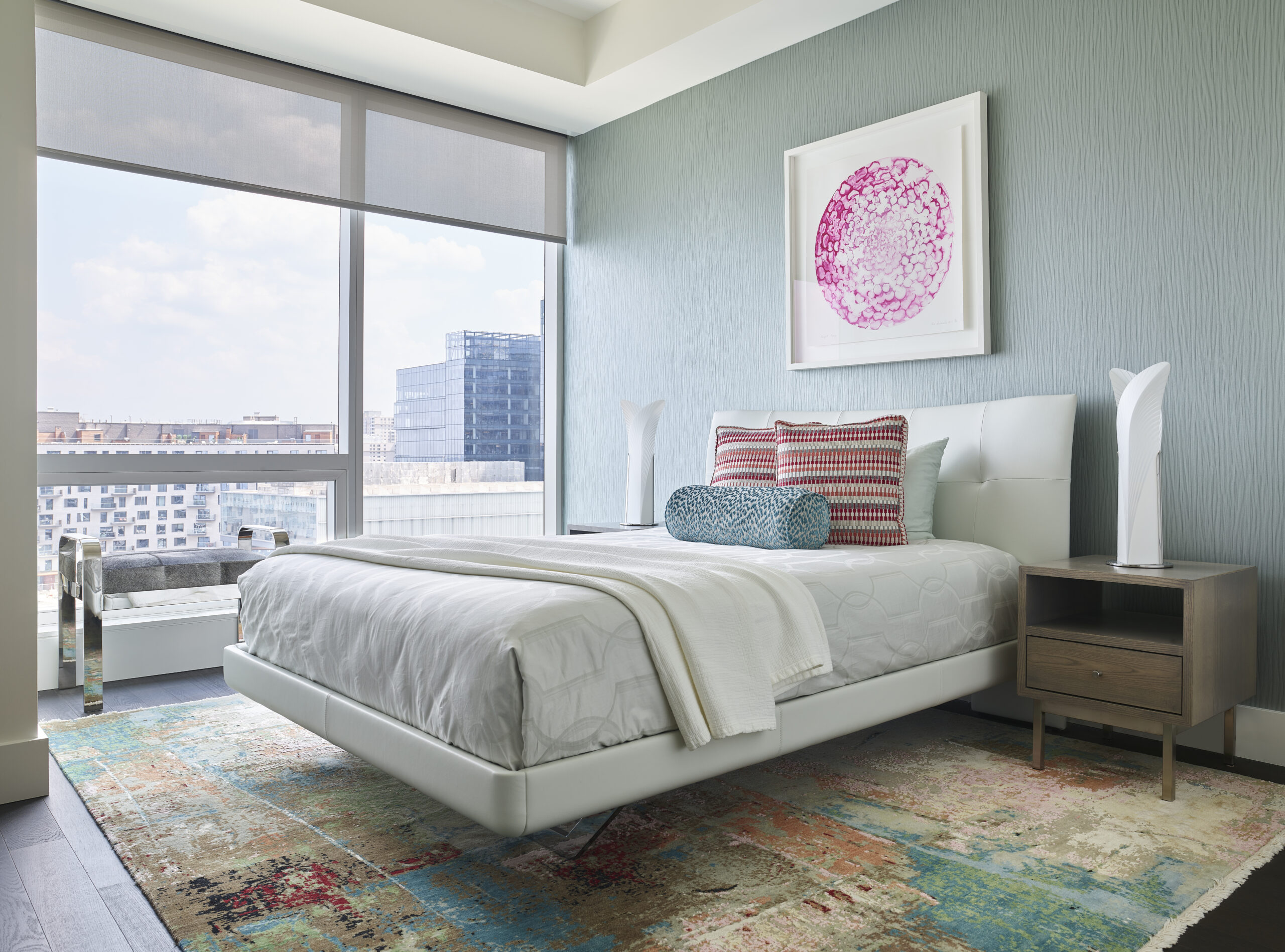 Modern bedroom with a large window, white platform bed, colorful rug, two bedside tables with lamps, decorative pillows, and abstract art on the wall—perfect for those seeking Luxury Interior Design.