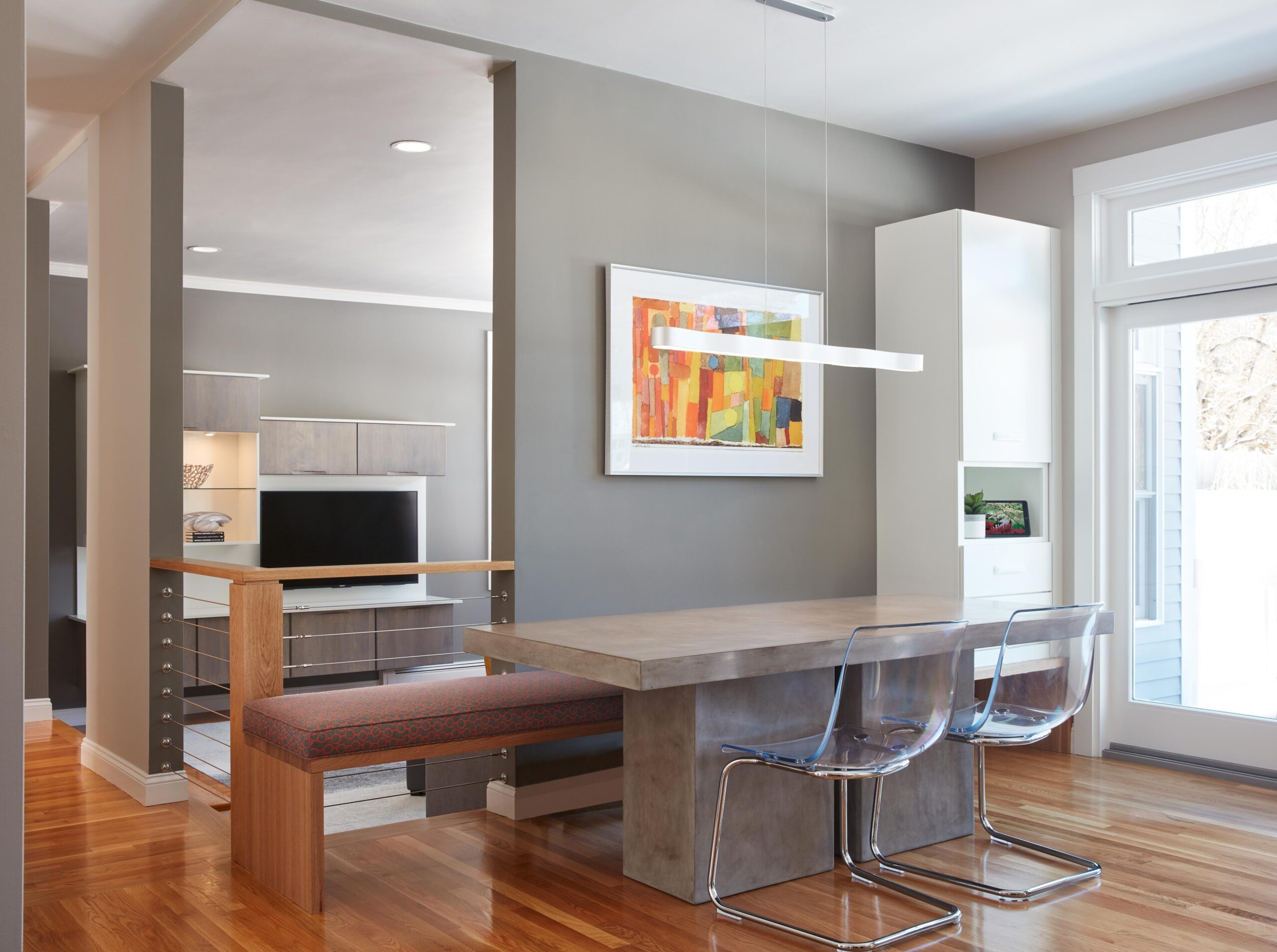 Modern dining area with a concrete table, clear chairs, built-in bench, abstract art on the wall, and large window letting in natural light—perfect for a stylish living space.