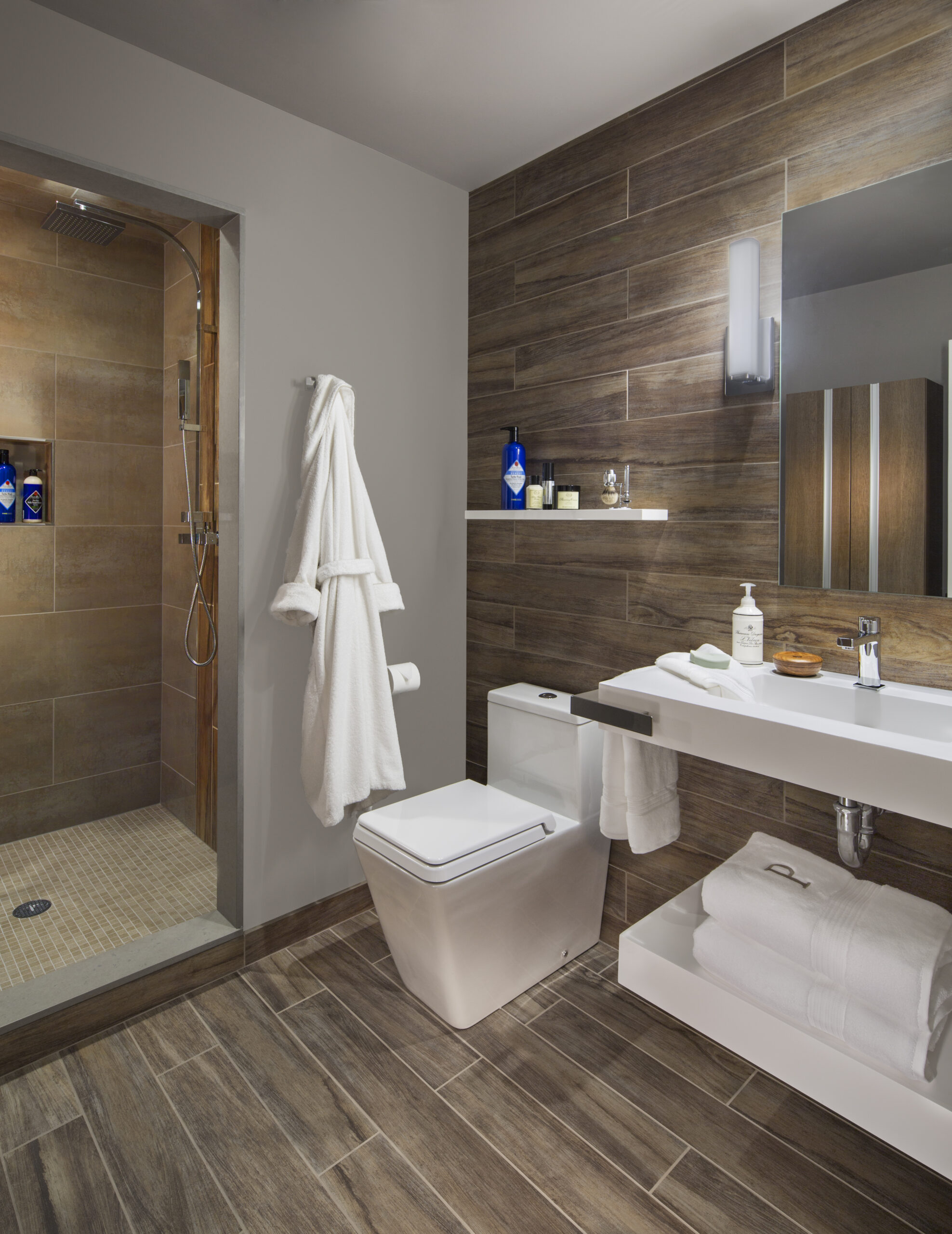 Modern bathroom with wood-look tile, a walk-in shower, white sink, wall-mounted toilet, robe on a hook, and toiletries—showcasing luxury interior design by an expert interior designer in Massachusetts.