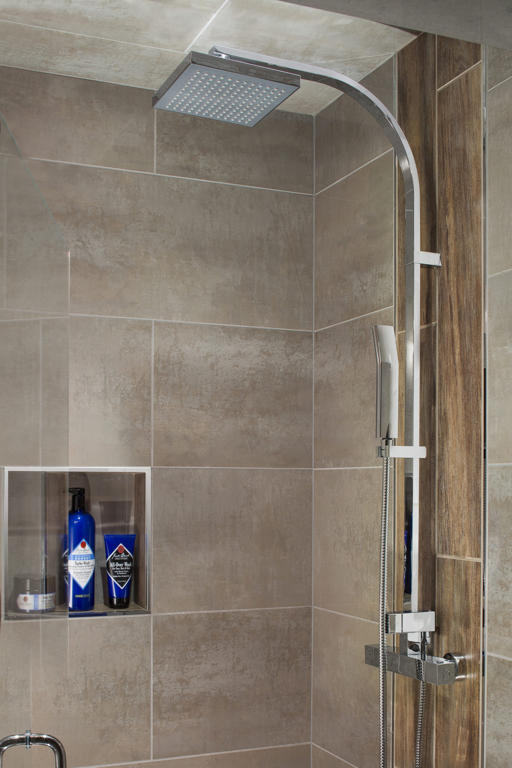Modern shower with a square overhead rainfall showerhead, a handheld attachment, and various products on a built-in shelf against tiled walls—perfect inspiration for those seeking luxury interior design by an interior designer in Massachusetts.