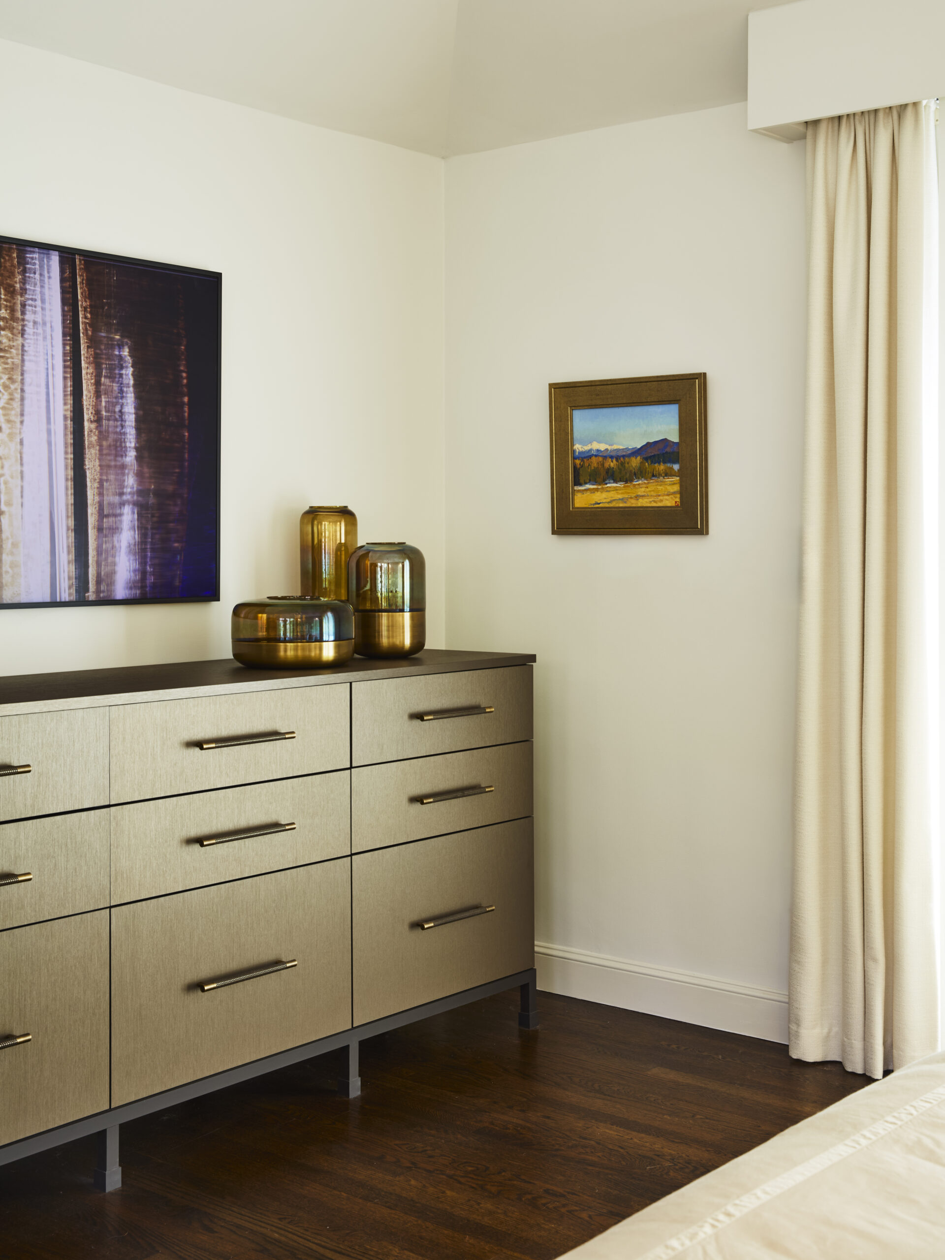A modern dresser with metallic finish stands against a white wall, topped with decorative glass vases—two framed artworks hang nearby, reflecting the touch of luxury interior design by an expert interior designer from Massachusetts.