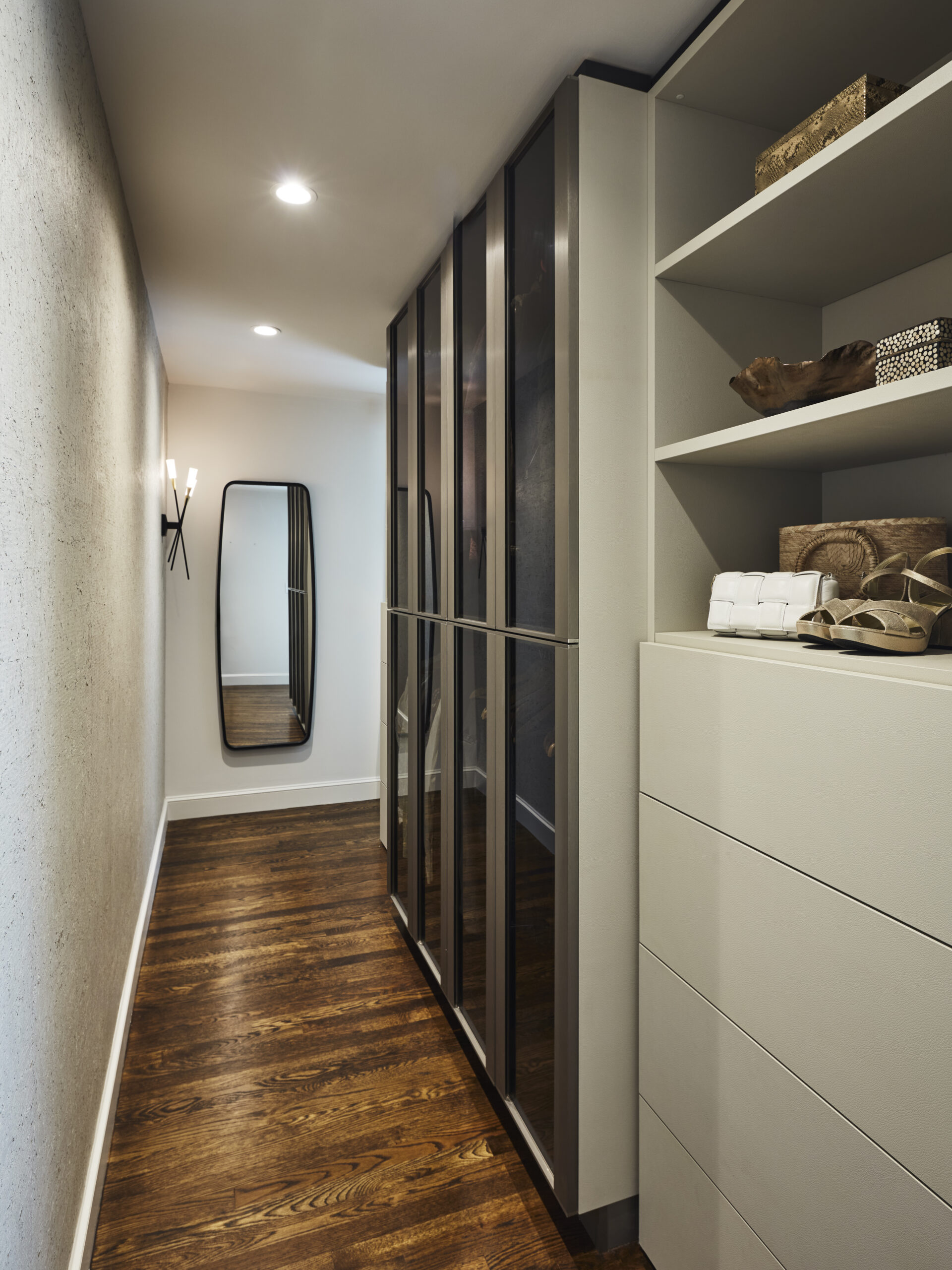 A modern walk-in closet with wood flooring, glass-front wardrobes, open shelving, and a wall mirror at the end of the hallway showcases luxury interior design at its finest.
