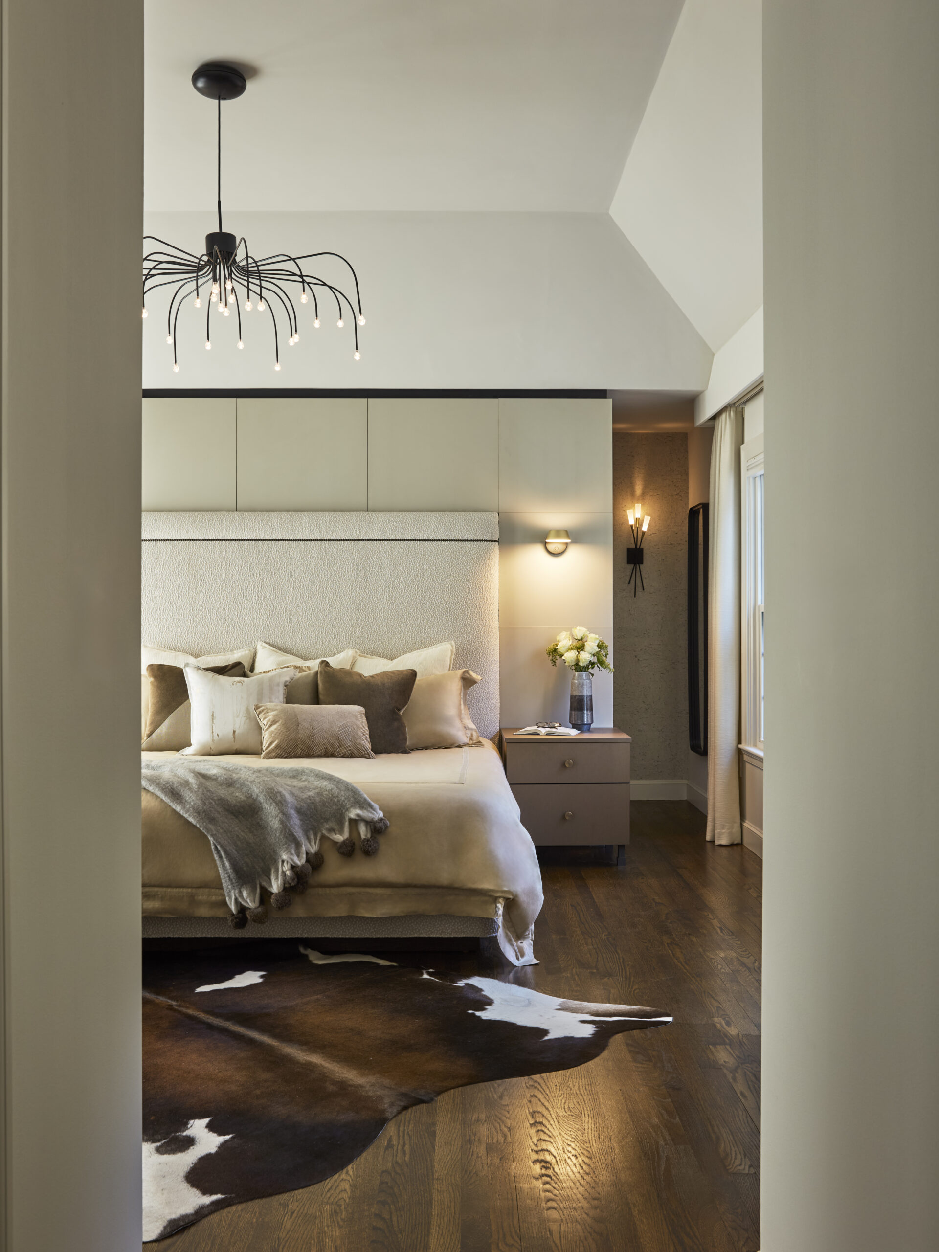 Modern bedroom with beige bedding, cowhide rug, nightstands, decorative pillows, and a contemporary chandelier—showcasing luxury interior design by an expert interior designer in Massachusetts—softly lit and viewed from the doorway.