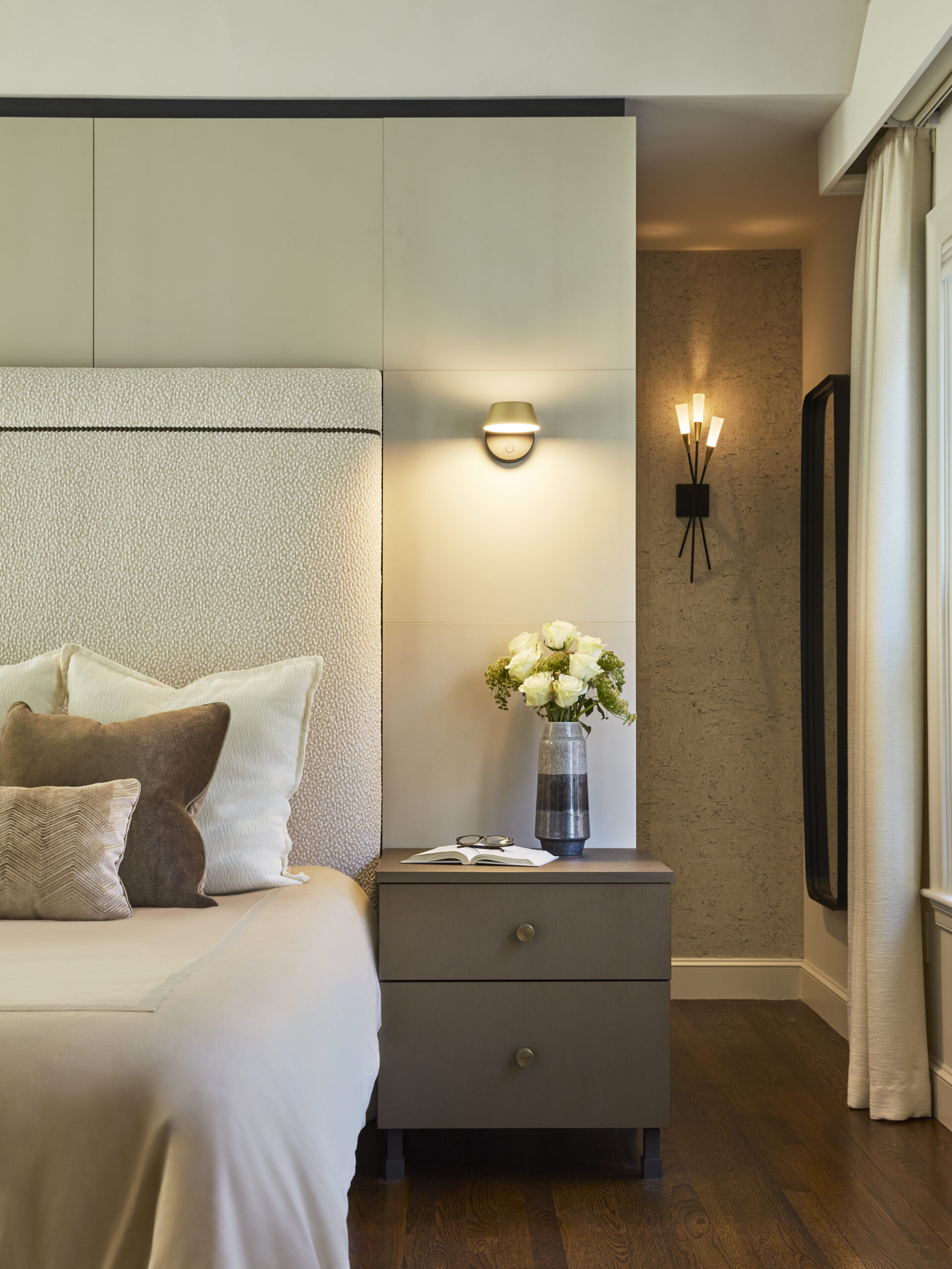 A modern bedroom by an Interior Designer in Massachusetts features a bed with beige linens and pillows, a nightstand with a vase of white flowers, and wall sconces—showcasing luxury interior design against neutral walls and a wood floor.
