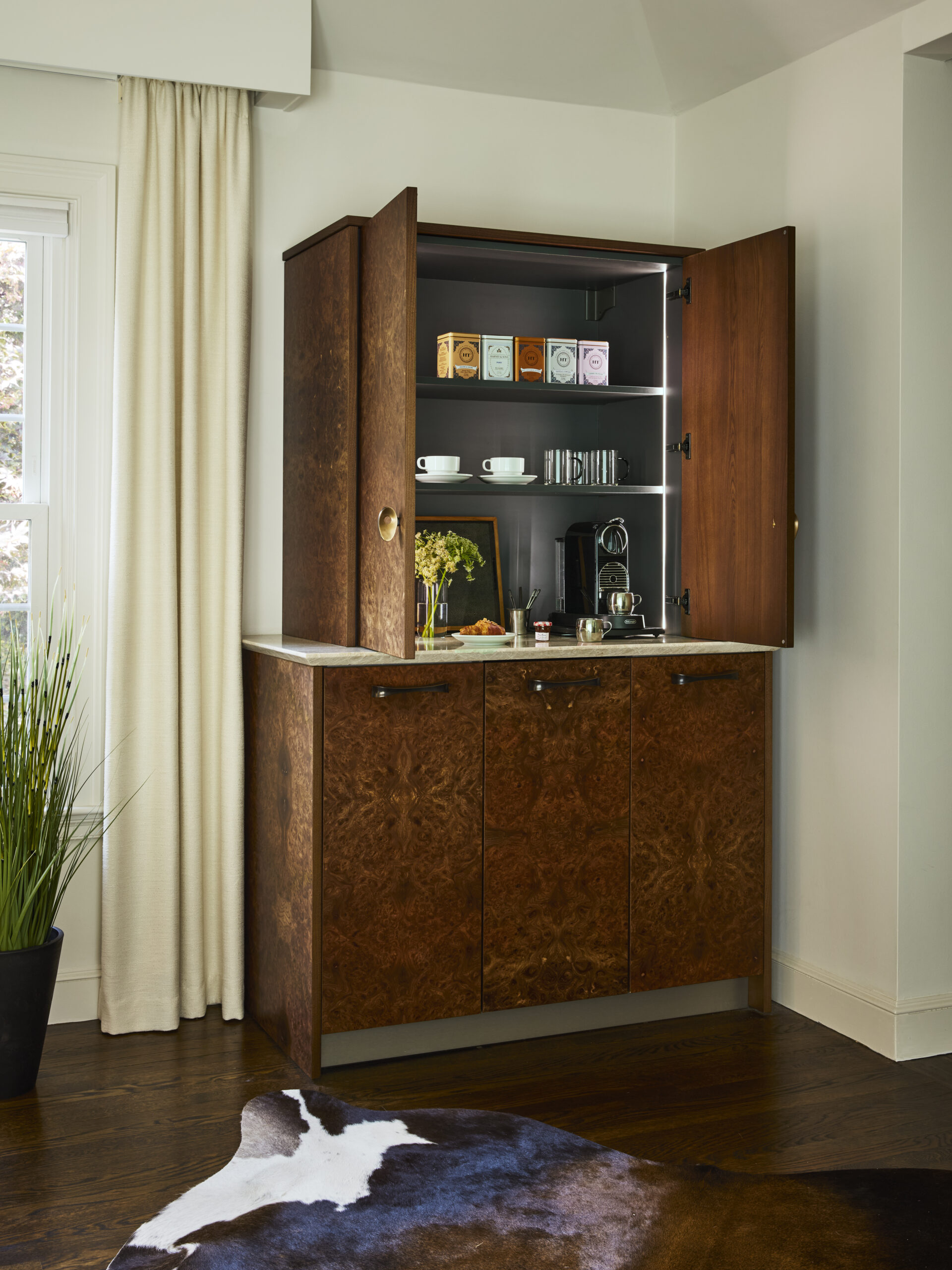 A wooden cabinet with open doors reveals a luxury coffee and tea station, including cups, a coffee machine, tea boxes, and glassware on shelves—an elegant touch inspired by luxury interior design, next to a window with white curtains.