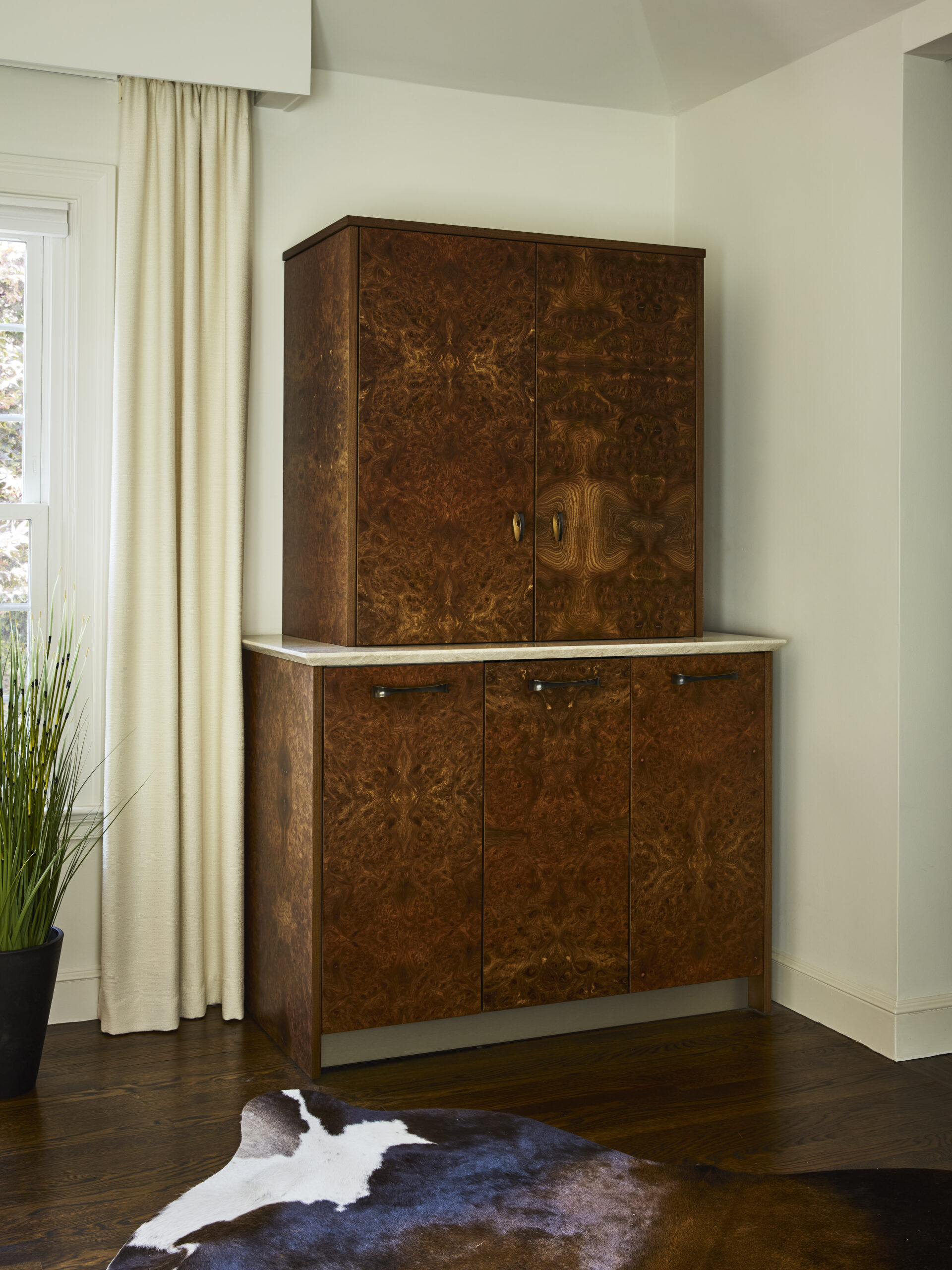A wooden cabinet with a marble top stands against a white wall next to a window with cream curtains and a potted plant; a cowhide rug on the dark wood floor adds to the luxury interior design crafted by an Interior Designer in Massachusetts.