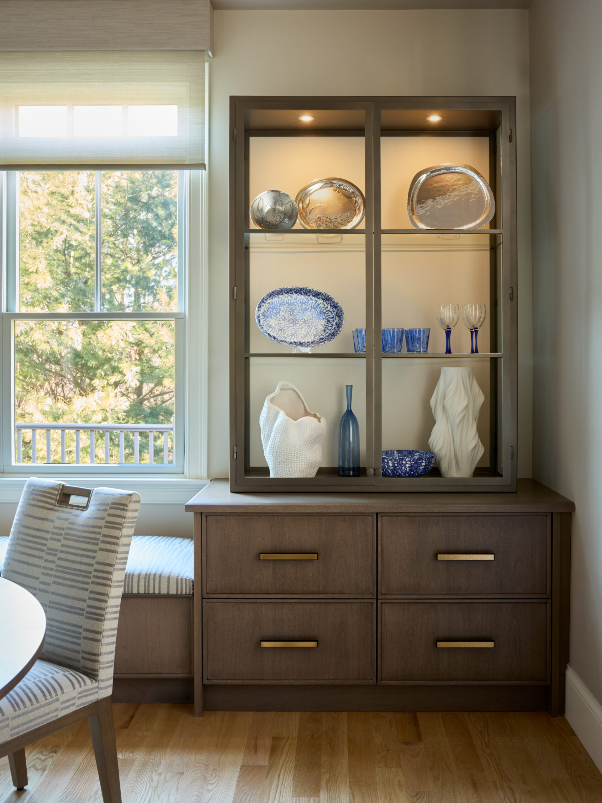 A wooden cabinet with drawers showcases blue and white decorative glassware, plates, and vases on shelves; sunlight from the window highlights the refined details—a touch of luxury interior design often seen by an interior designer Massachusetts trusts.