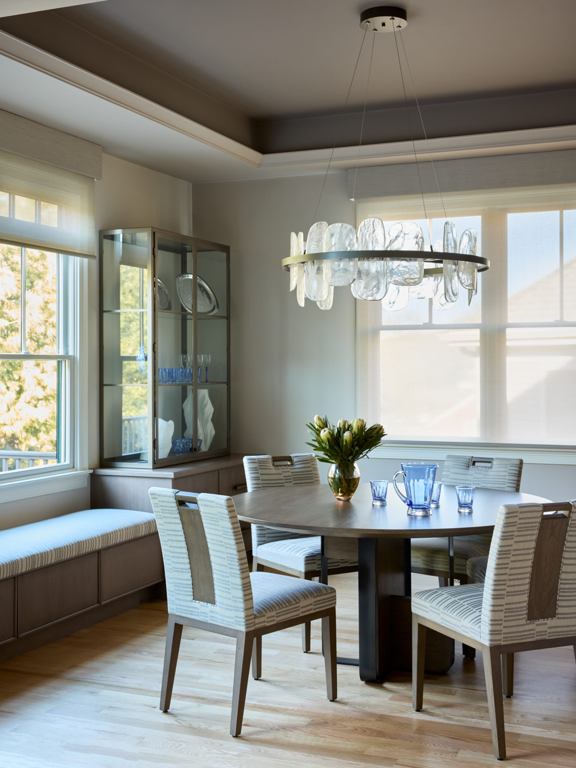 Modern dining room with a round table, four chairs, a glass chandelier, built-in bench seating, and a display cabinet with dishes by large windows—an inviting space that reflects the sophistication of Luxury Interior Design.