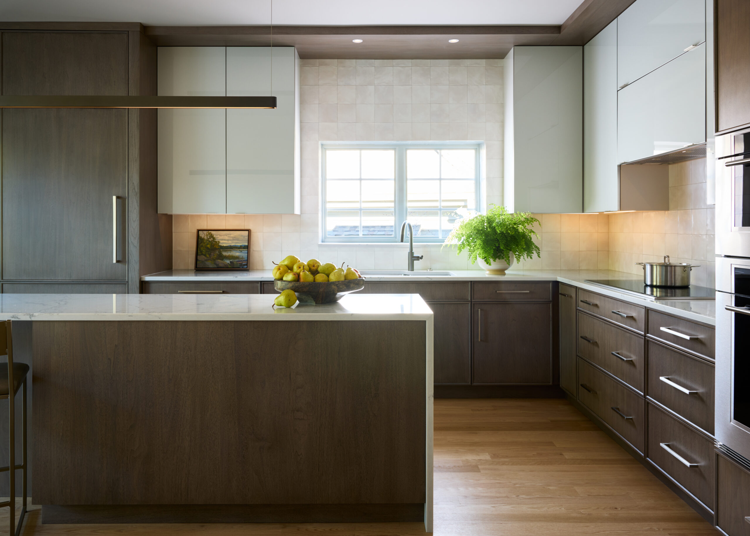 Modern kitchen with wood cabinets, white countertops, island with fruit bowl, potted plant by the sink, and natural light coming through a large window—an elegant touch of luxury interior design by an expert Interior Designer Massachusetts.
