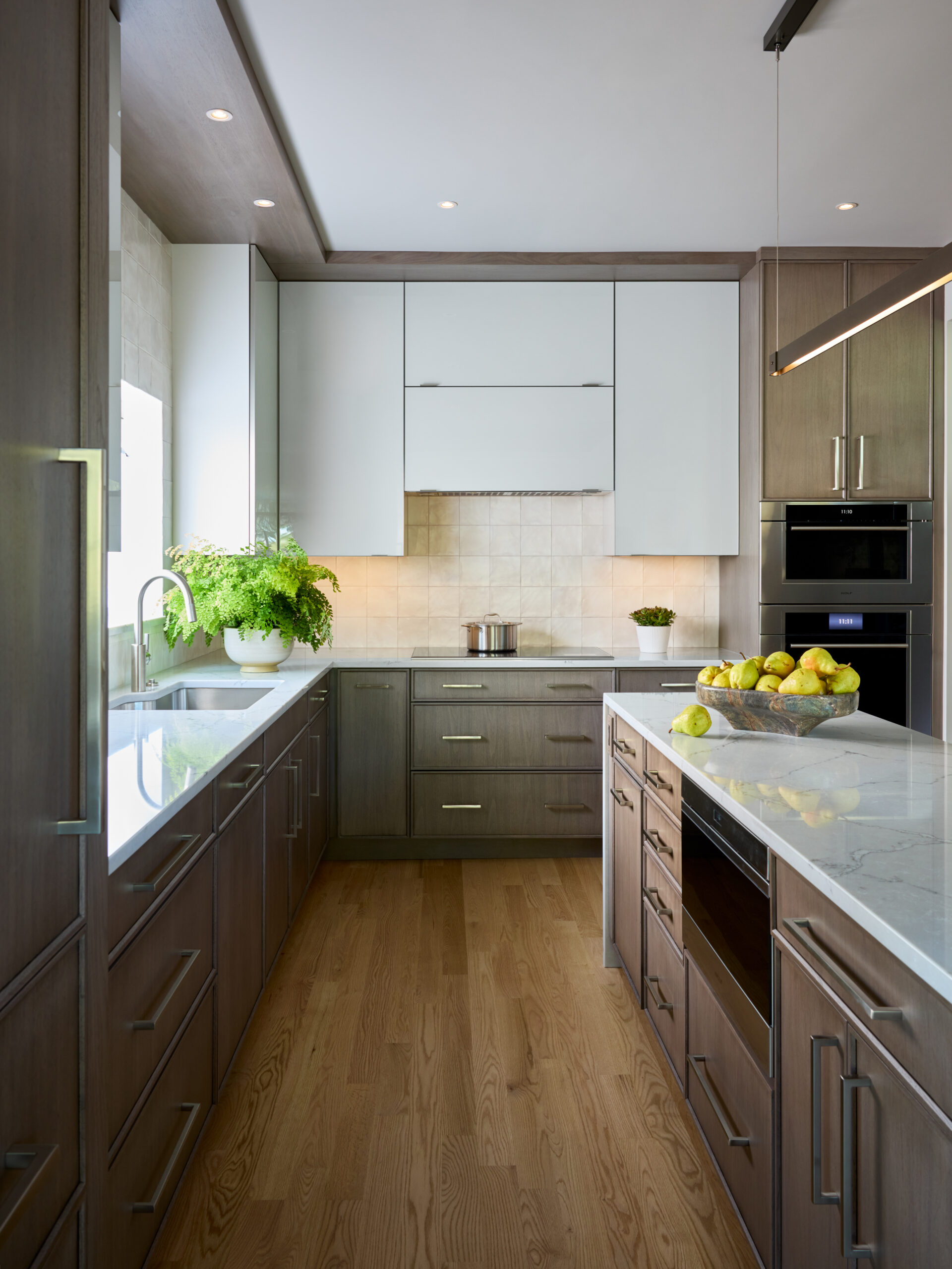Modern kitchen with wood cabinetry, white countertops, stainless steel appliances, potted plants, and a bowl of apples on the island. Designed by a luxury interior design expert in Massachusetts, natural light enters from a window by the sink.
