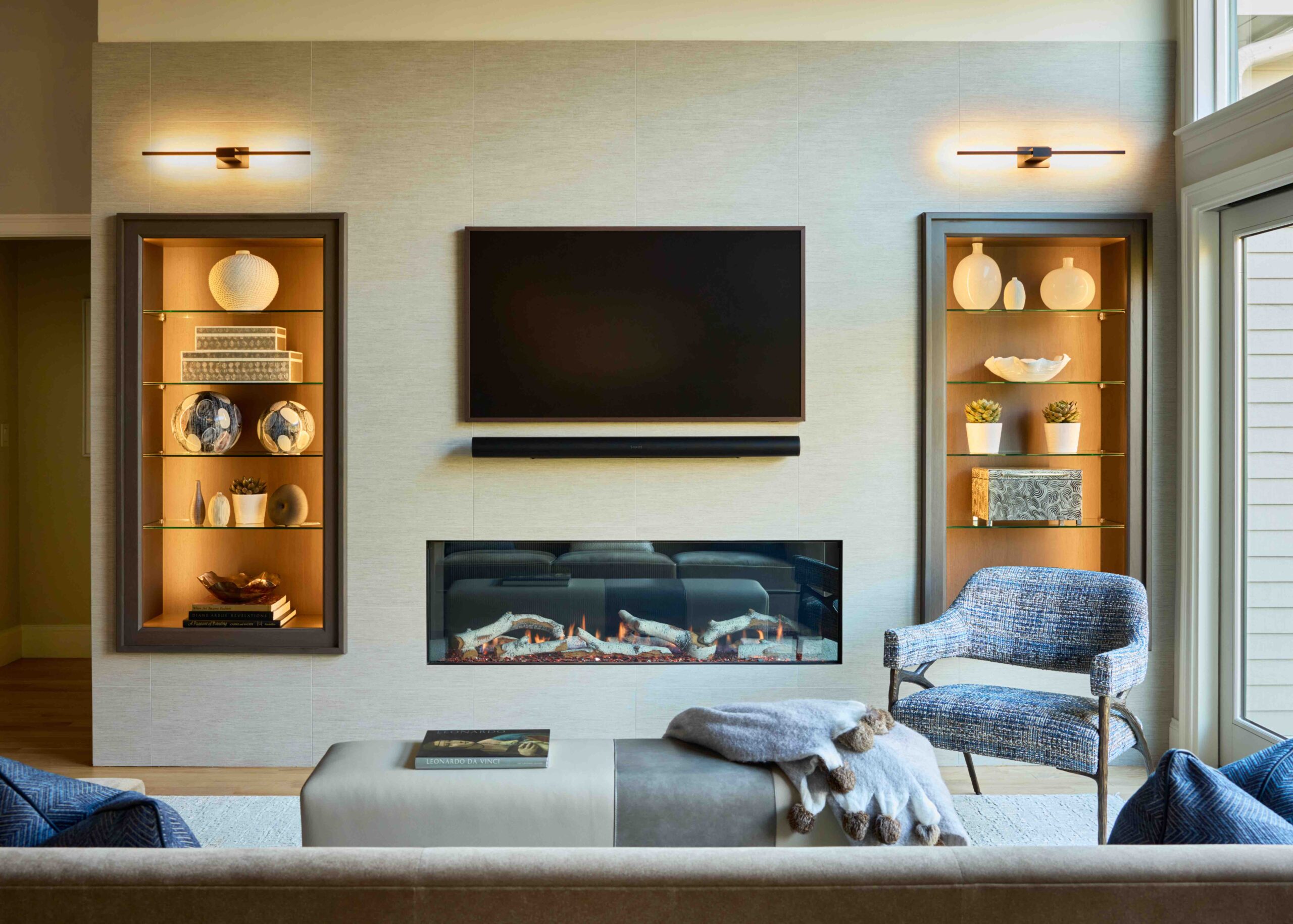 Modern living room featuring thoughtful interior design, with a wall-mounted TV above a linear fireplace, flanked by shelves displaying decorative items; armchair and ottoman in the foreground.