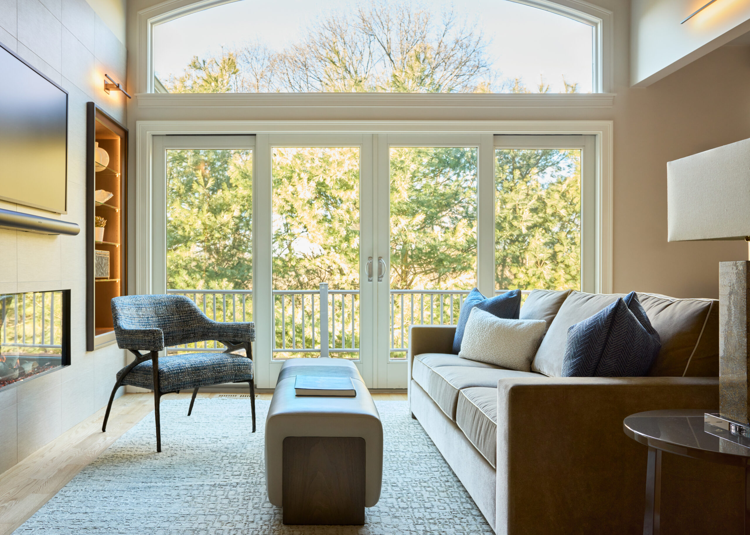 A modern living room with a sofa, armchair, and ottoman showcases luxury interior design, featuring large windows and a stunning view of trees outside.