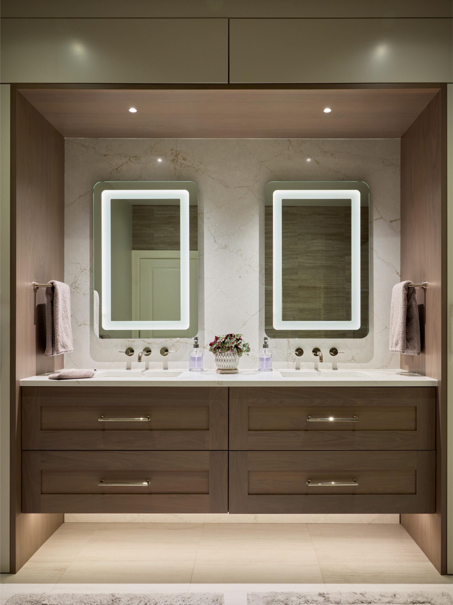 Modern bathroom with double sinks, light wood cabinetry, illuminated rectangular mirrors, marble backsplash, and towels hanging on both sides—showcasing the elegance achievable with expert Interior Design Services.