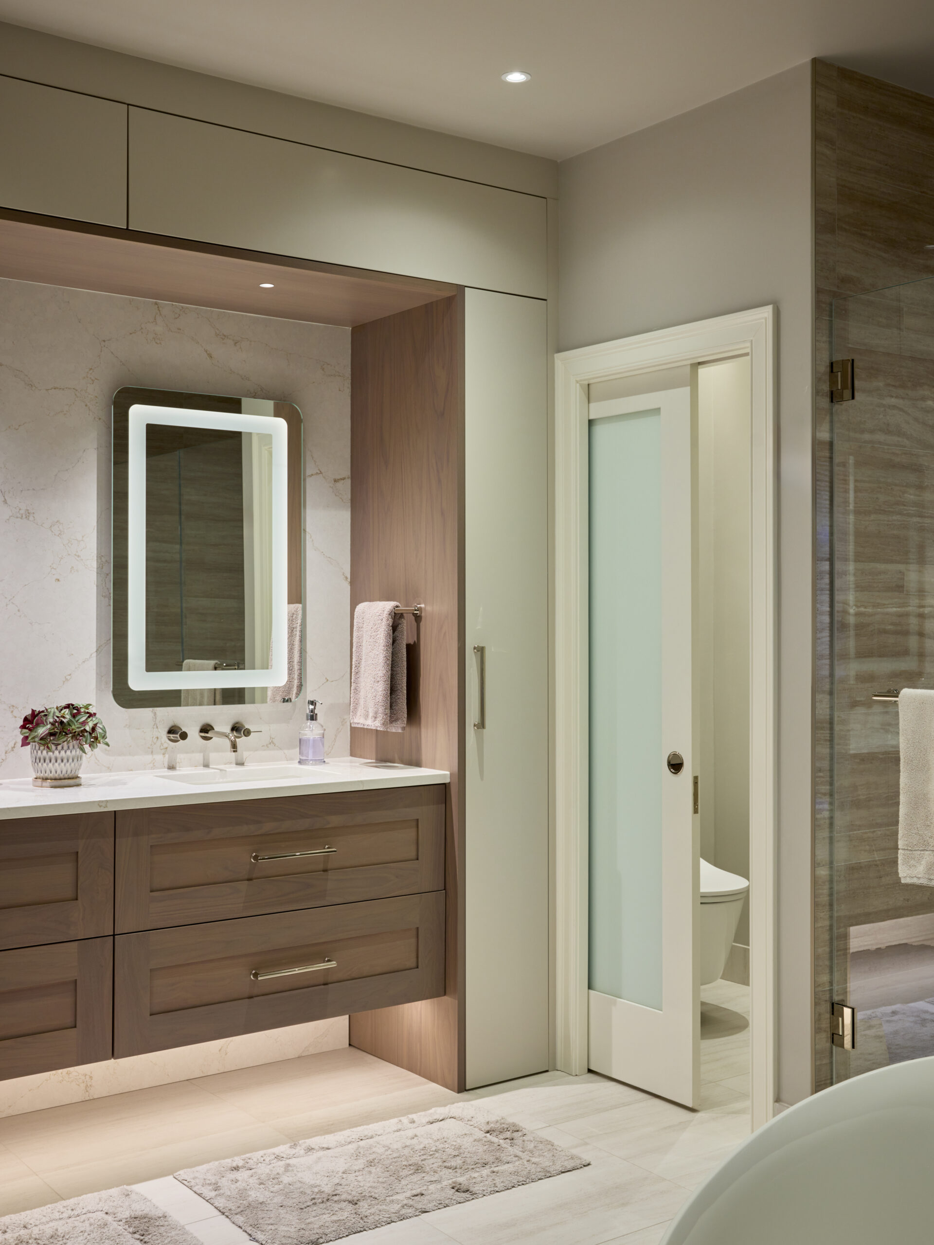 Modern bathroom with double sink vanity, illuminated mirror, marble backsplash, and sliding frosted glass door. Neutral tones reflect Luxury Interior Design. Towels and toiletries are neatly arranged for a sophisticated look.