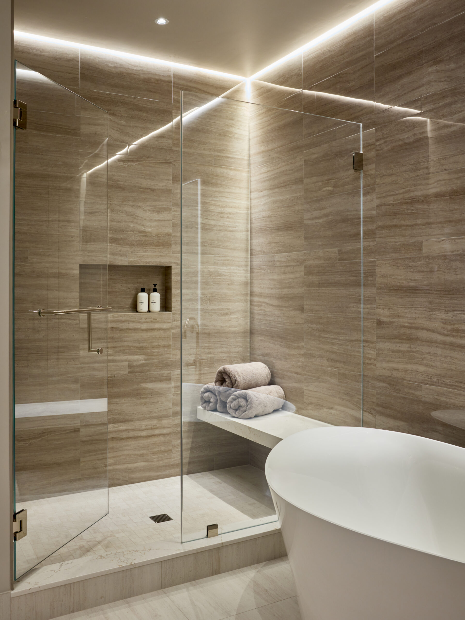 Modern bathroom with a glass-enclosed shower, built-in shelf holding toiletries, folded towels on a bench, and a white freestanding bathtub—an elegant example of Luxury Interior Design by an expert interior designer in Massachusetts.