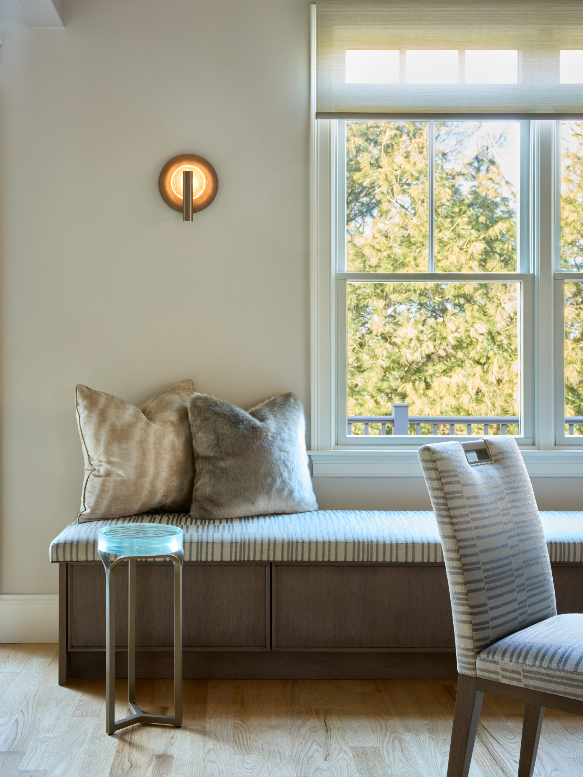 A window seat with a striped cushion and two plush pillows sits next to a window; reflecting Luxury Interior Design, a modern wall sconce and small side table are nearby as sunlight filters through the trees outside.