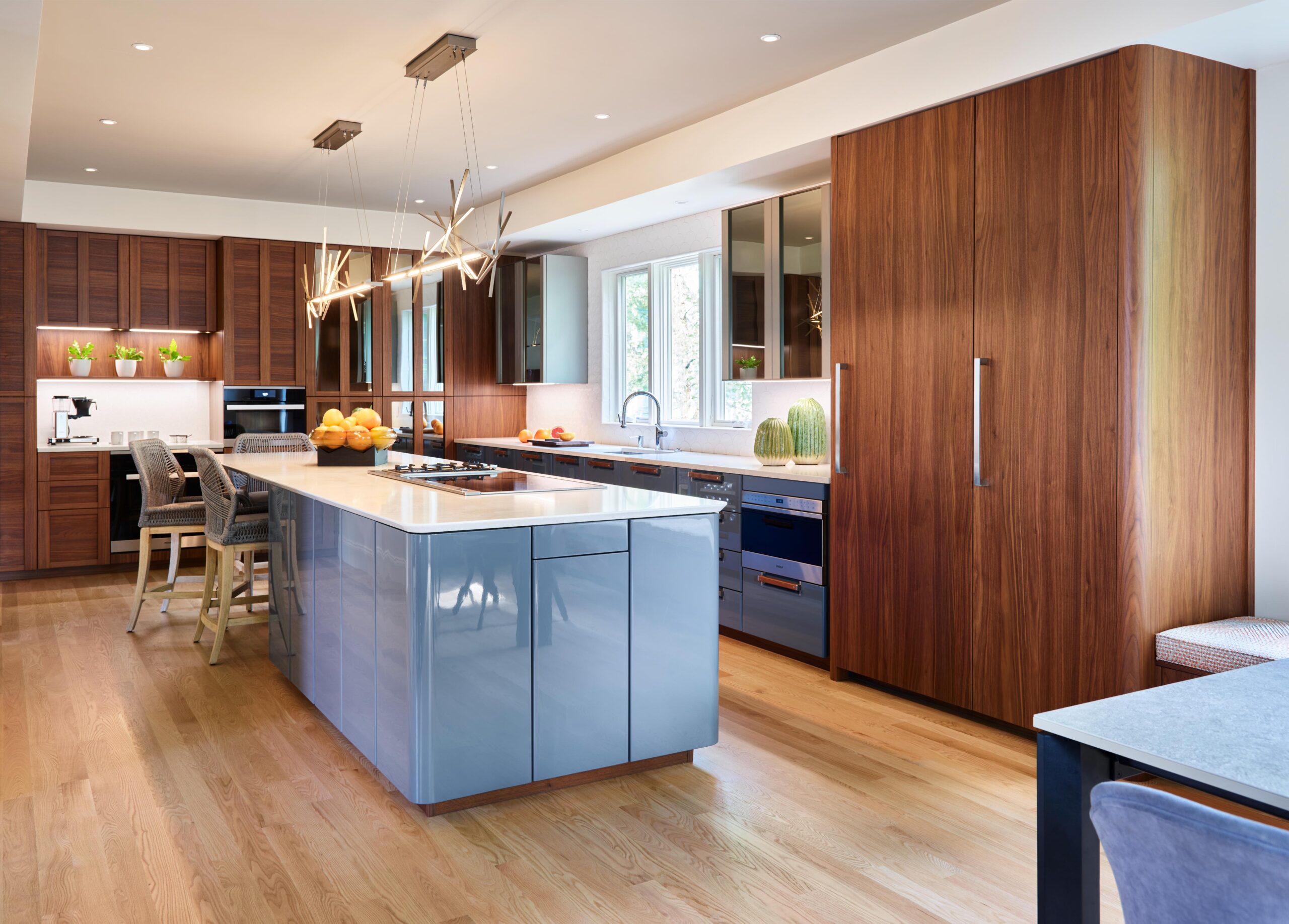 Modern kitchen with a glossy blue island, wooden cabinets, built-in appliances, pendant lights, and light wood flooring. Fruit bowls and decor on the countertops showcase thoughtful touches from expert interior design services.